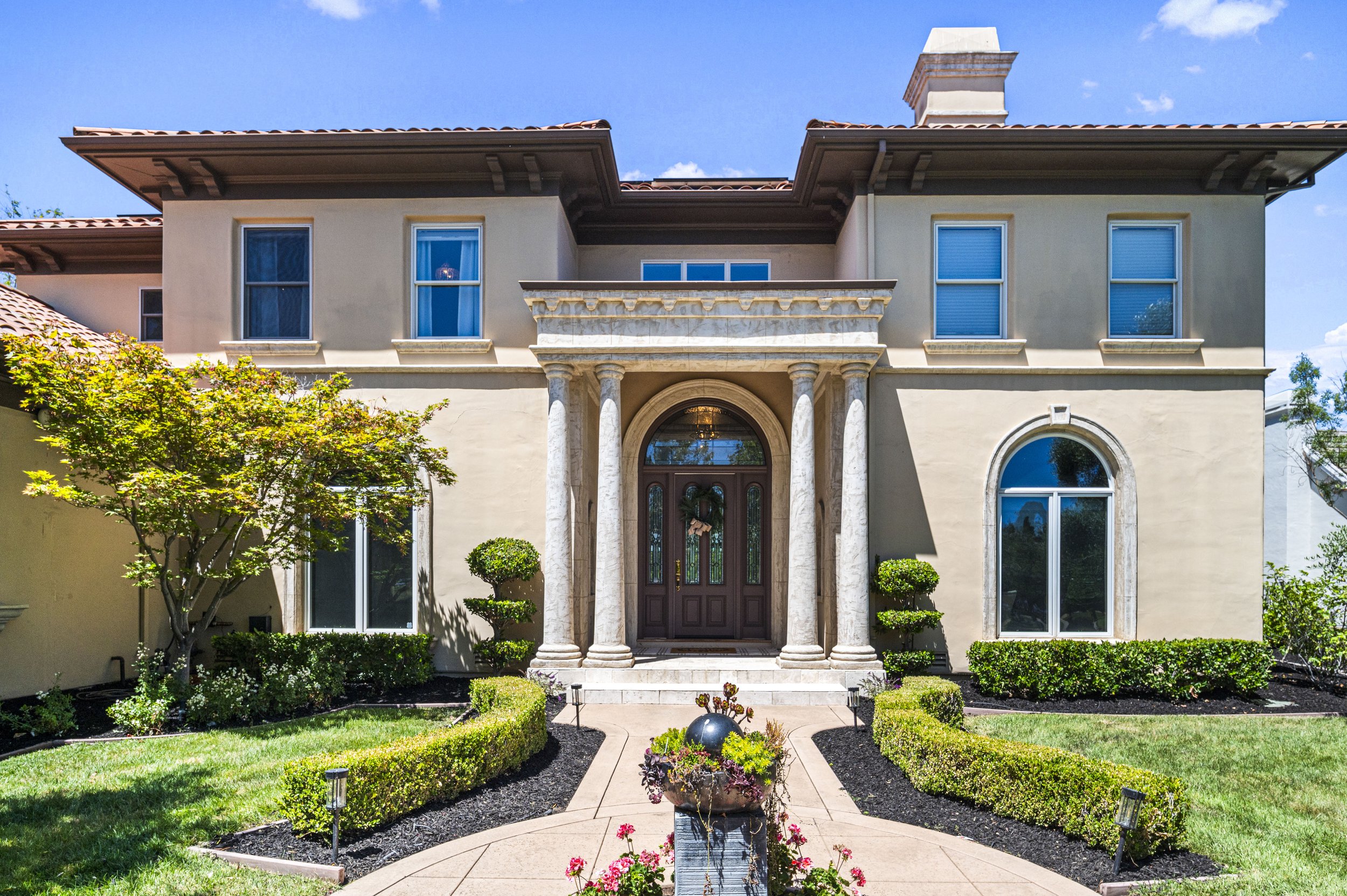 Front view of a two-story house with a landscaped front yard, trees, bushes, a walkway leading to the front door, and sunlit clear blue sky.