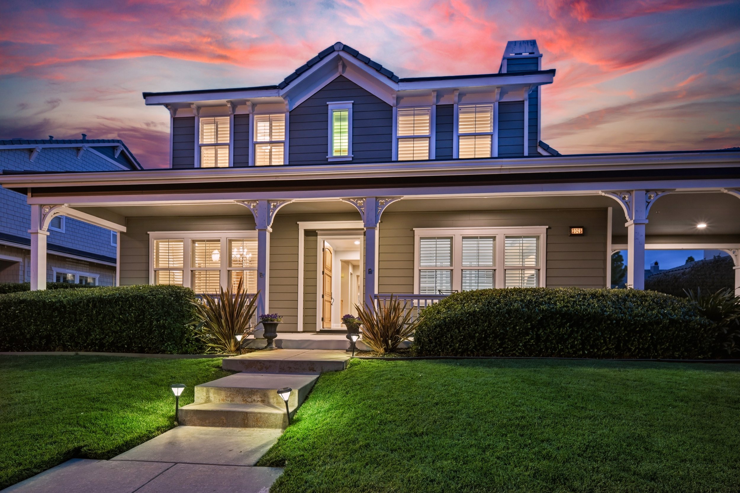 A two-story house at dusk with warm interior lighting, gray exterior, front porch, decorated garden, and a colorful sunset sky.