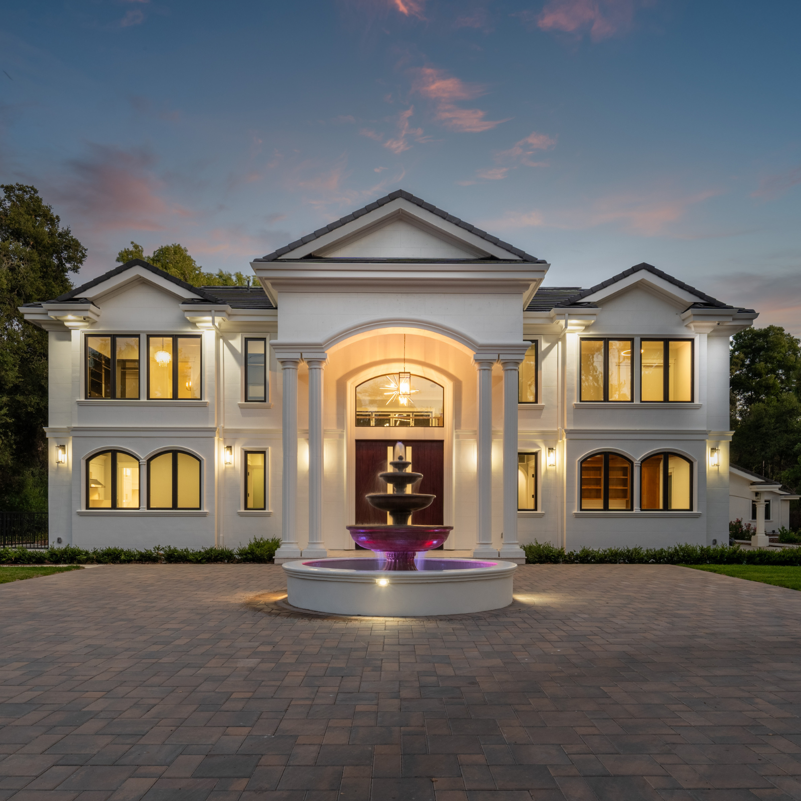 Large white mansion with multiple windows, front entrance with columns, and a fountain with purple lighting in front, during dusk.