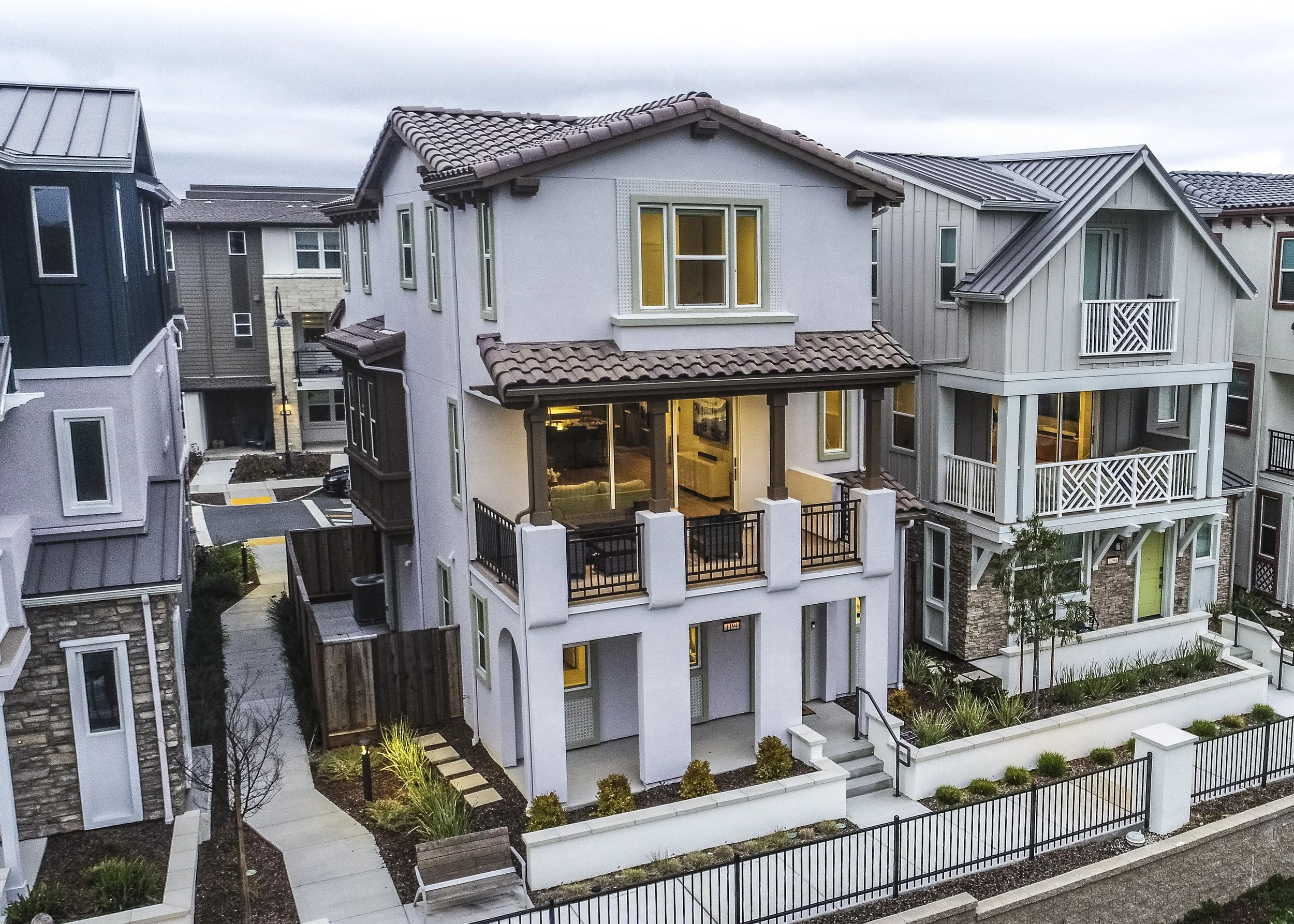Multi-story modern residential houses with tile roofs, large windows, and balconies in a neighborhood, with outdoor lighting and landscaped yards.