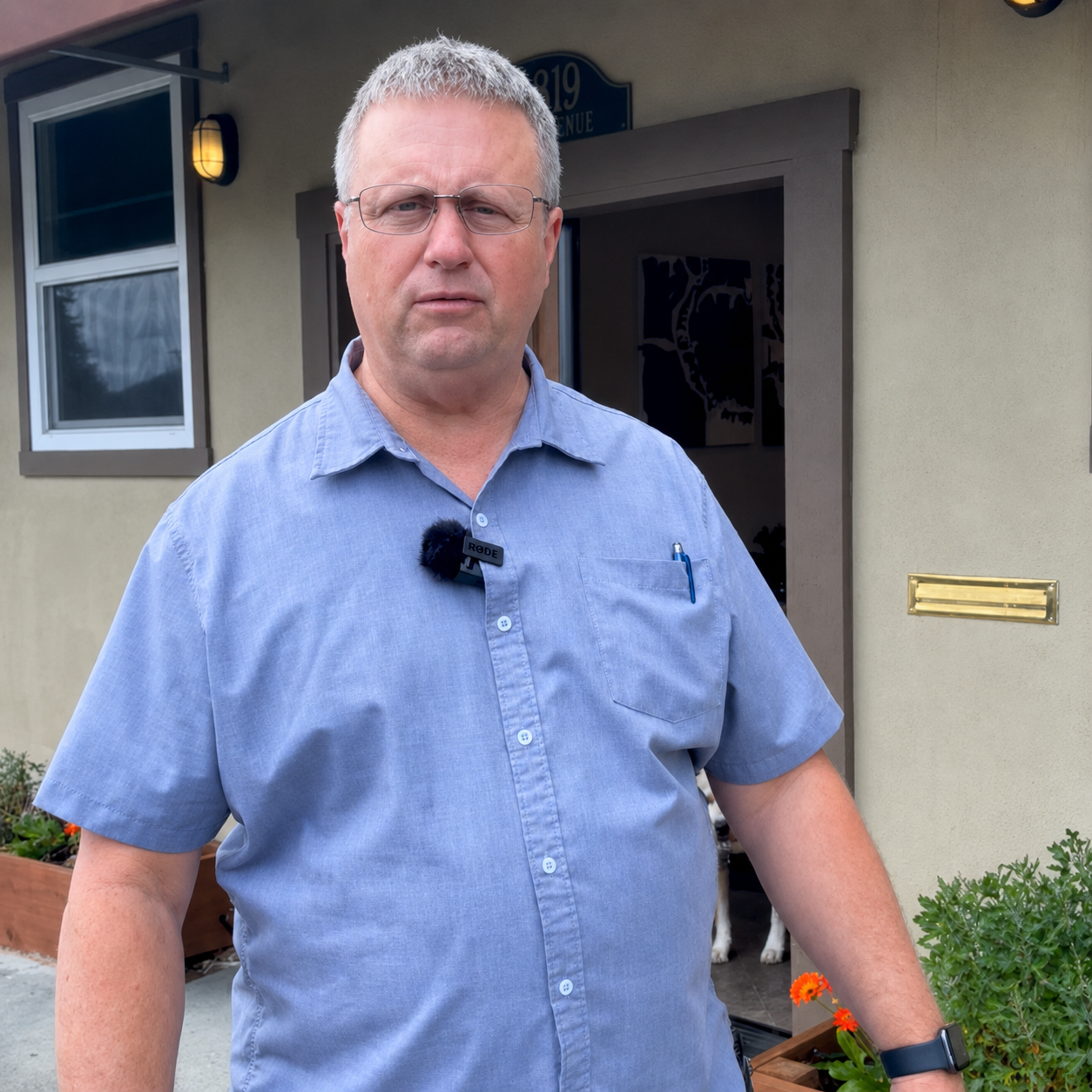 A middle-aged man with gray hair, glasses, wearing a blue button-up shirt, standing outdoors near a house with a window and a door, with plants and flowers nearby.