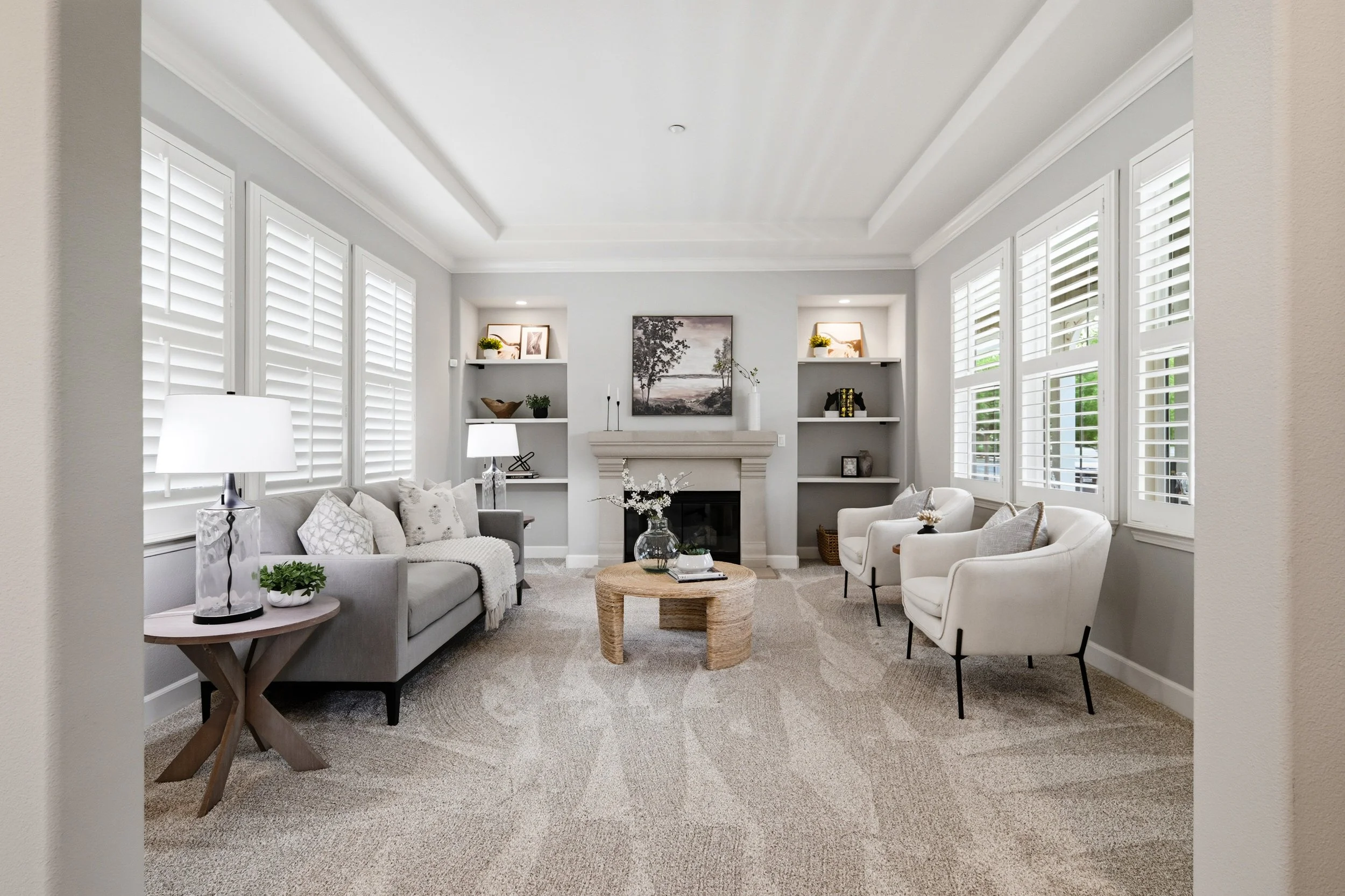 Living room with white walls, multiple windows with white shutters, a fireplace, sofas, armchairs, and decorative items such as framed pictures, lamps, plants, and sculptures.
