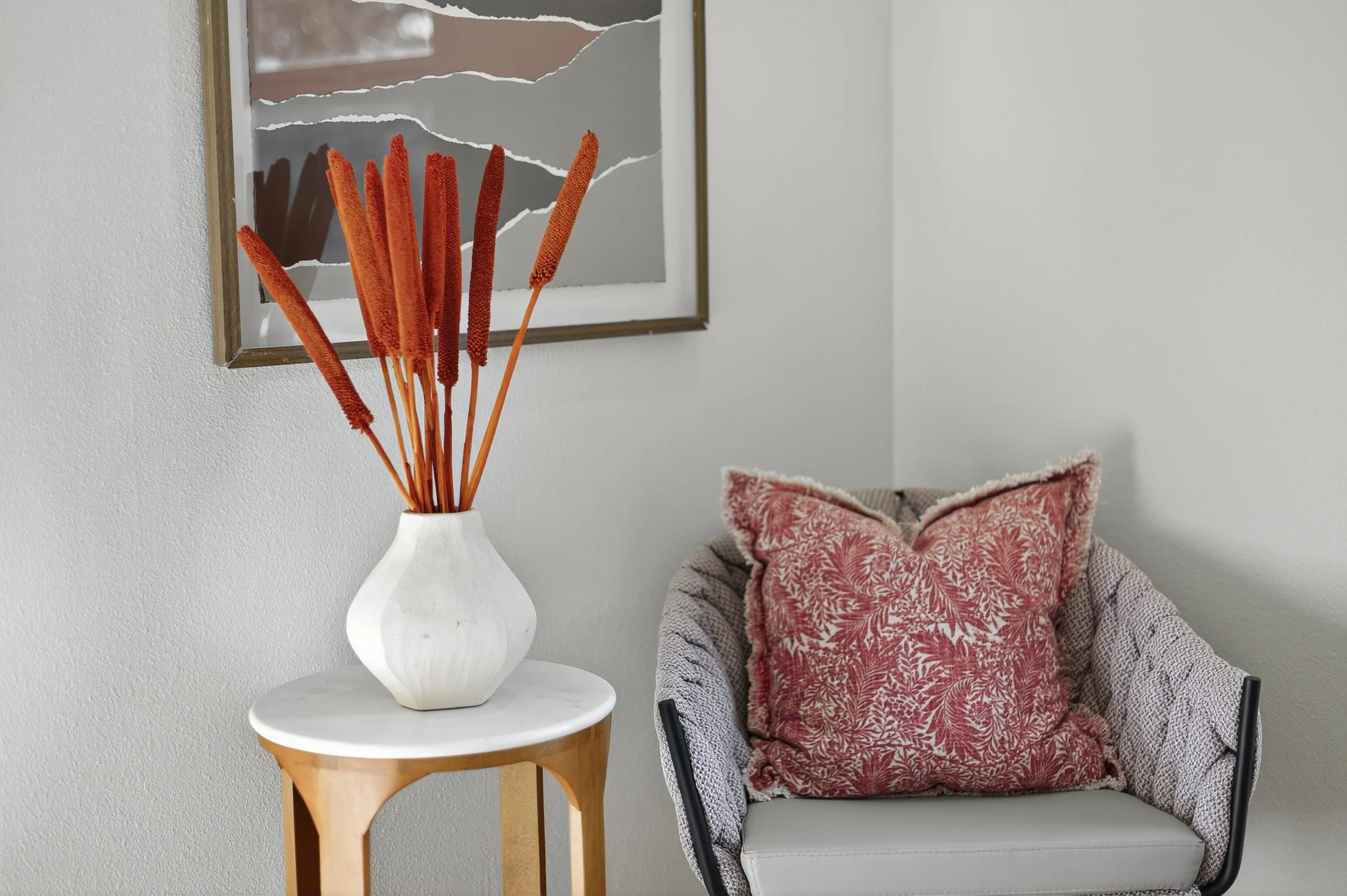 A white vase with orange dried flowers placed on a small round wooden table. In the background, there is a beige armchair with a patterned red pillow and a gray blanket draped over the armrest, set against a plain white wall with a torn mirror hanging on it.