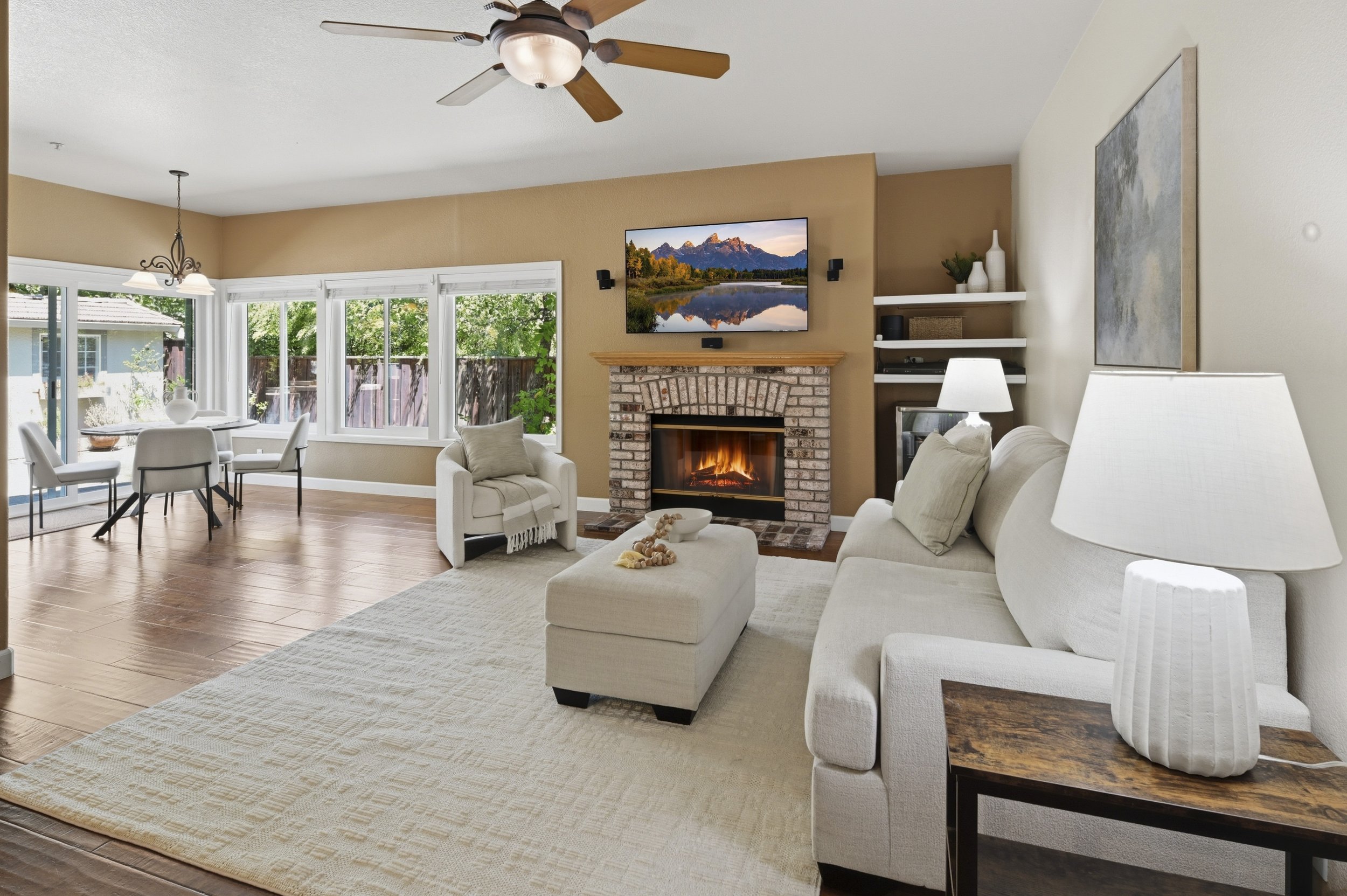 Bright living room with white sofa, armchair, brick fireplace, flat-screen TV, ceiling fan, large windows, and hardwood floors.