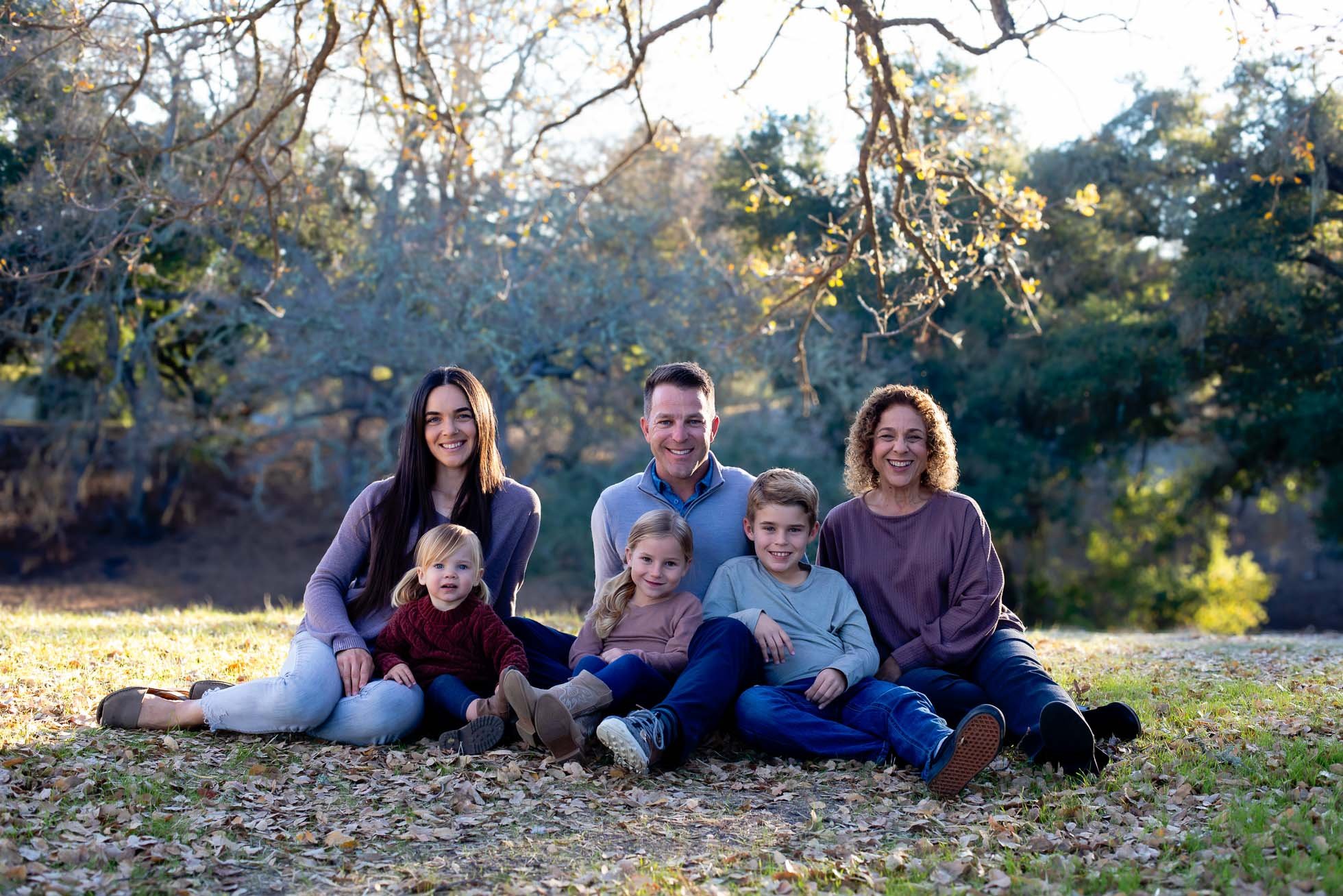 A multigenerational family sitting on fallen leaves outdoors in a park in autumn, smiling at the camera.