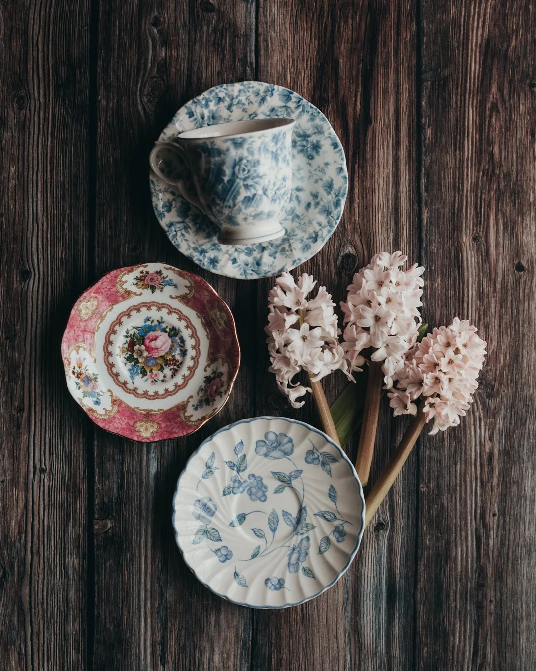 A wooden table holding a blue floral teacup and saucer, a pink floral plate, a white plate with blue floral designs, and three pink hyacinth flowers.