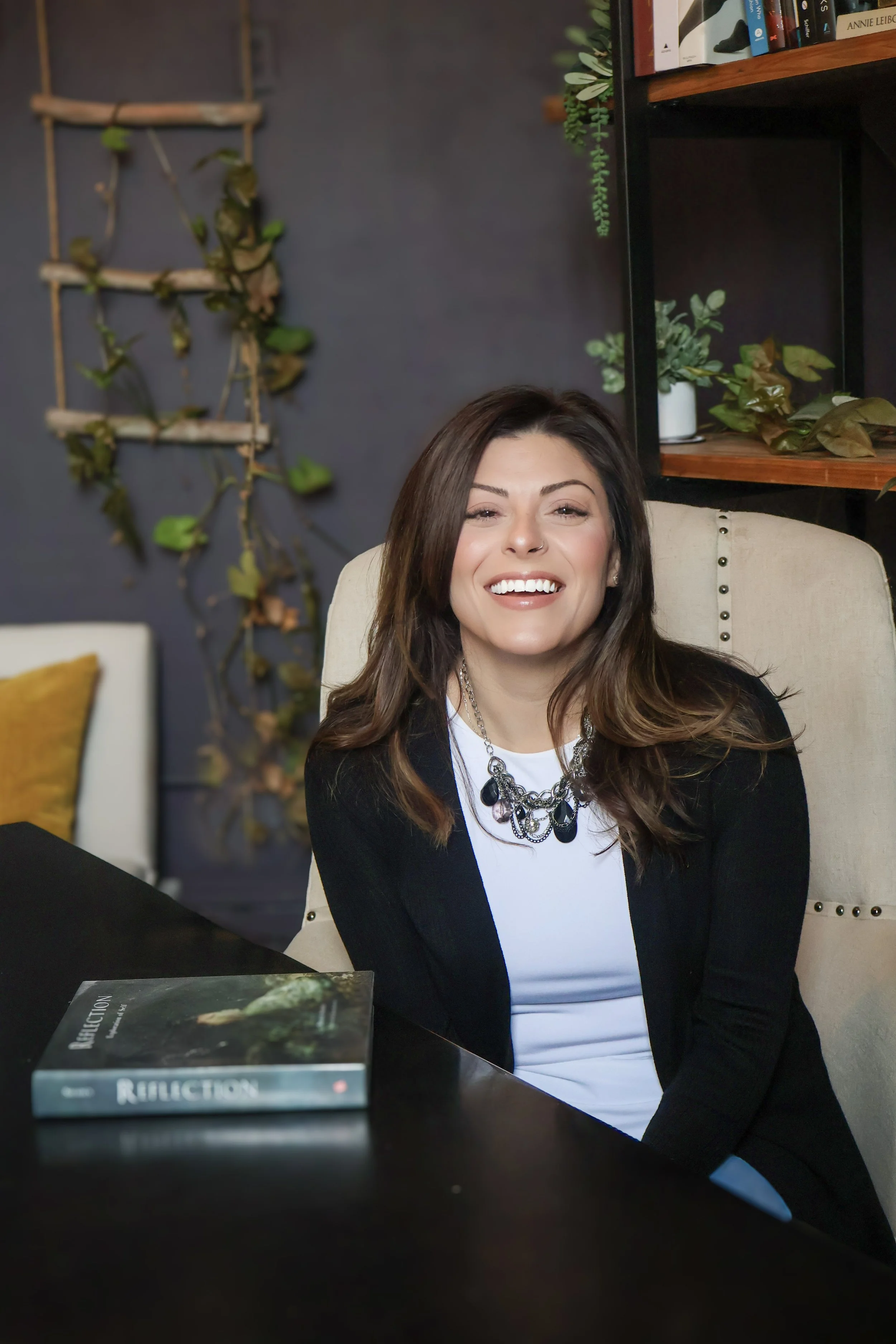 A woman with brown hair sitting at a black table, smiling and wearing a black jacket and a white top with a statement necklace, in a room decorated with plants and a bookshelf.