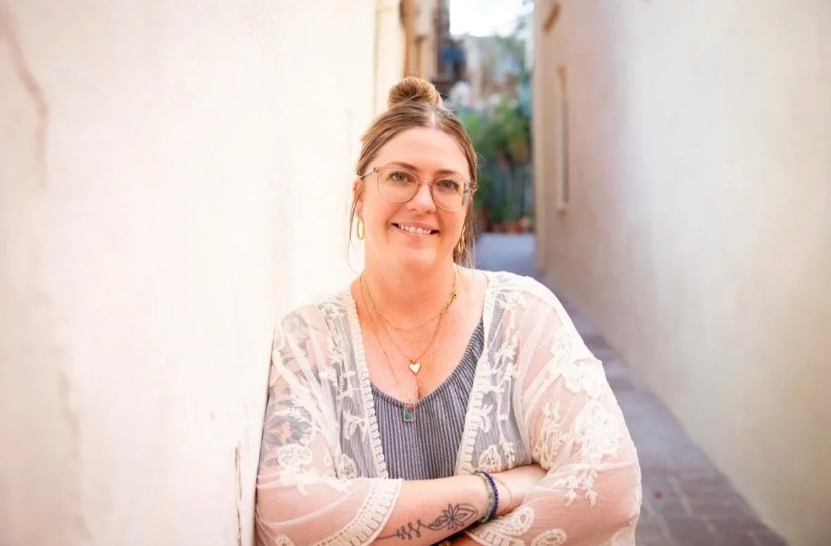 Smiling woman with glasses, tattoo on arm, wearing layered jewelry, standing in an alleyway with light-colored walls.