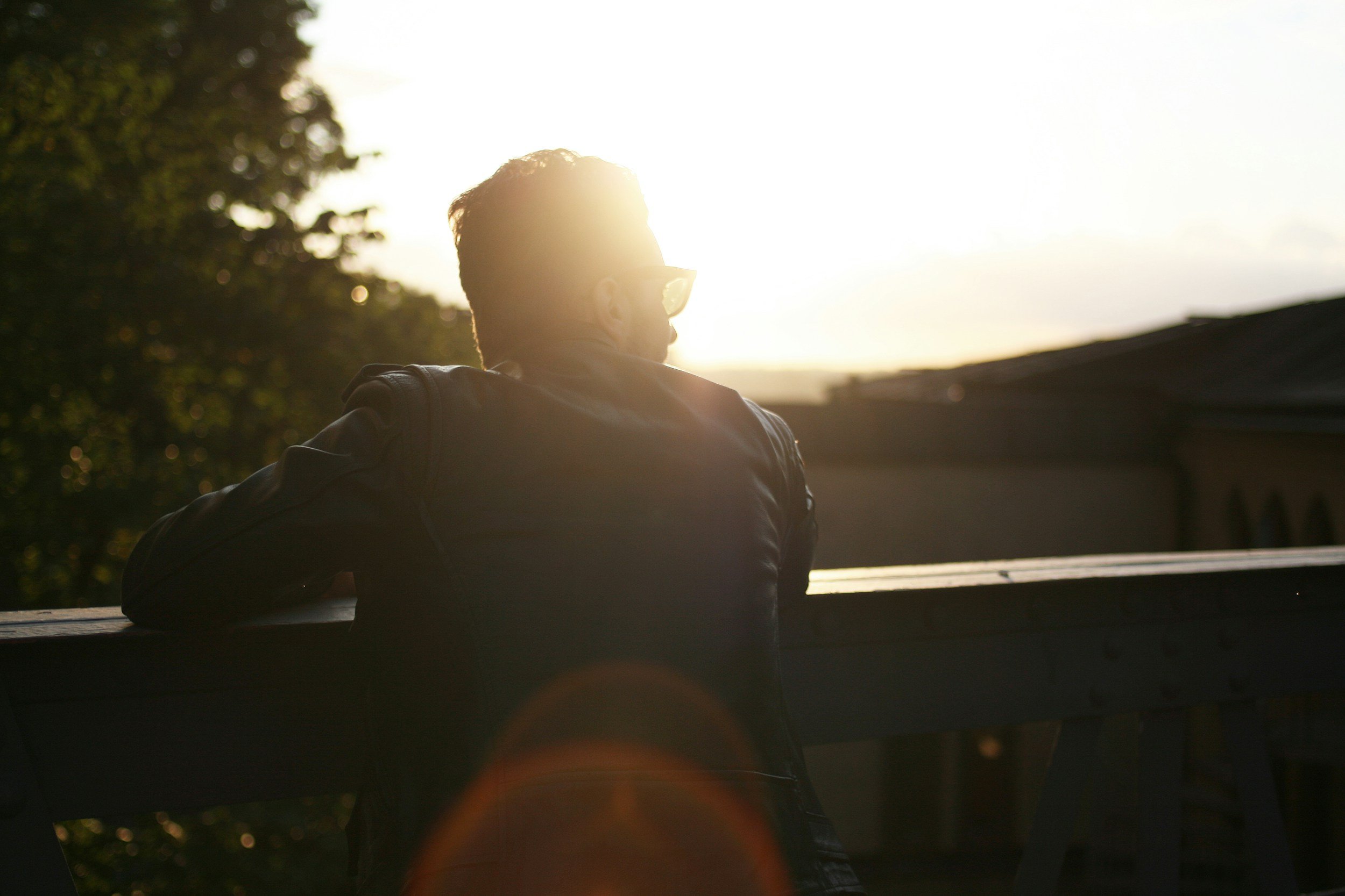 A person wearing sunglasses and a black jacket, leaning on a wooden railing and looking towards the sunset or sunrise.