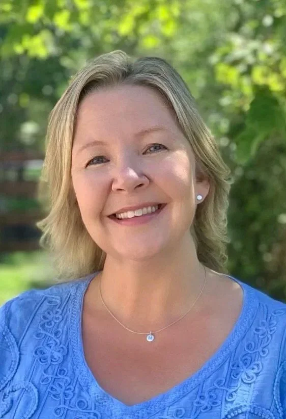 A smiling woman with short blonde hair wearing a blue embroidered top and a delicate necklace outdoors with green trees in the background.