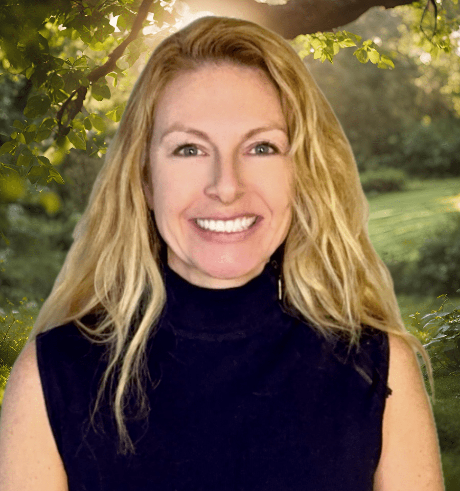 A woman with long blonde hair and a black sleeveless turtleneck smiling outdoors with greenery and trees in the background.