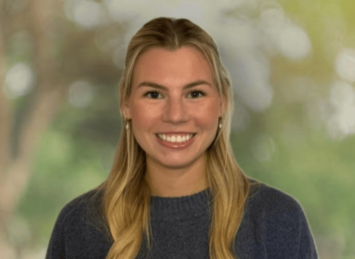 Portrait of a smiling young woman with blonde hair, wearing a dark gray sweater and earrings, against a blurred outdoor background.