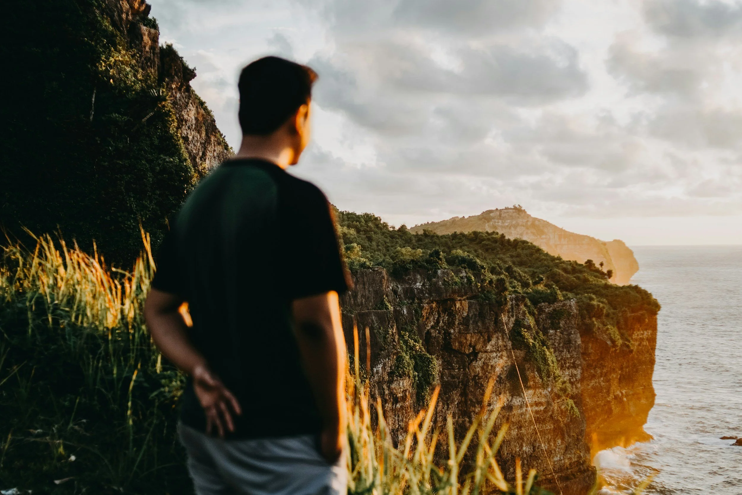 A man standing on a grassy cliff overlooking the ocean and cliffs at sunset.