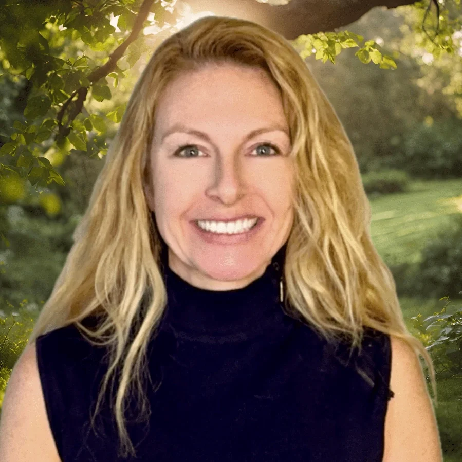 A woman with long wavy blonde hair smiling outdoors with trees and sunlight in the background.
