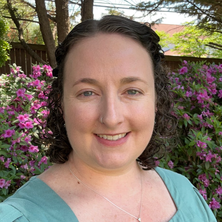 A woman with curly dark hair, light skin, and a nose piercing smiling outdoors, with a background of pink azalea flowers and trees.