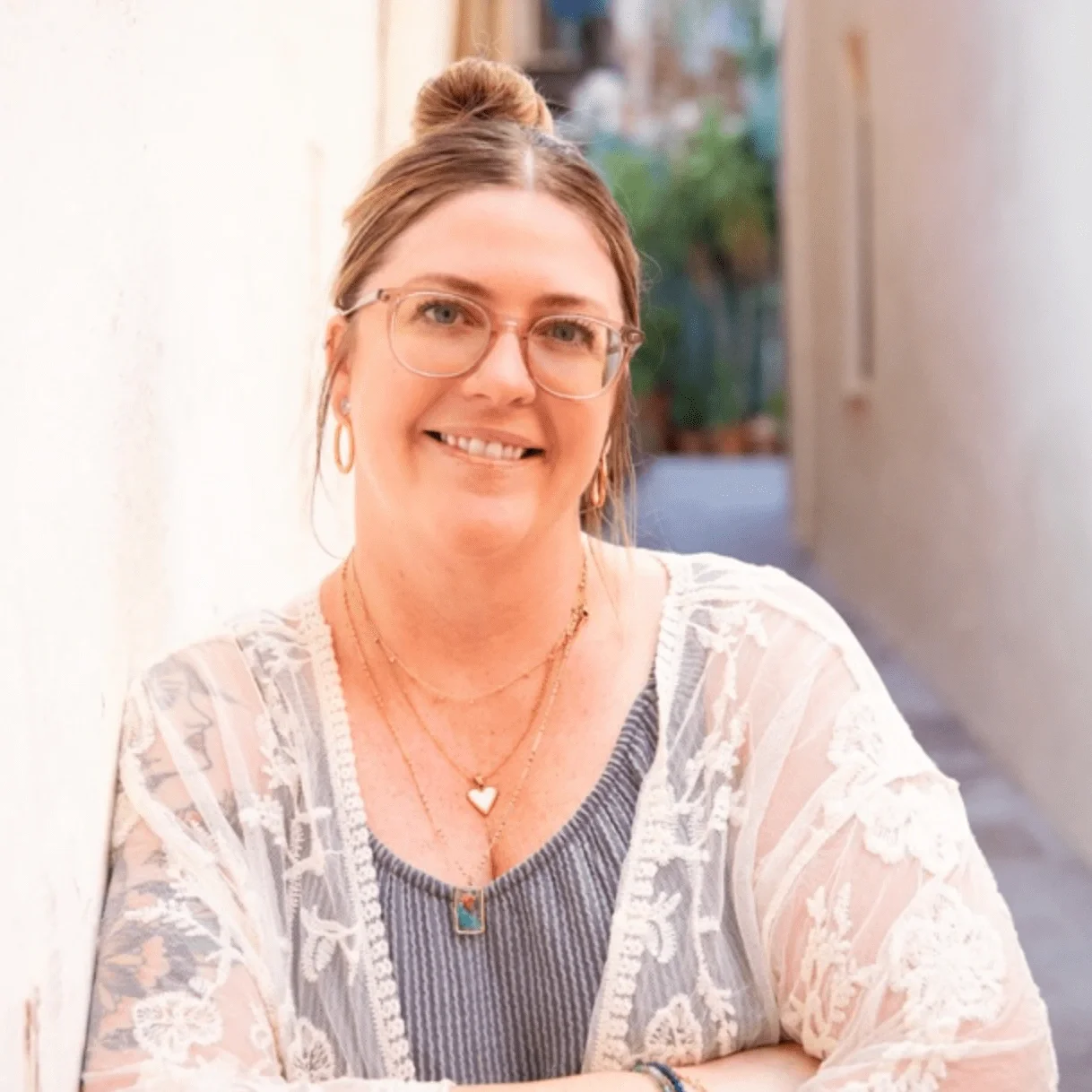 A smiling woman with glasses and earrings, wearing a white lace top and layered necklaces, standing outdoors in a narrow alleyway with greenery in the background.