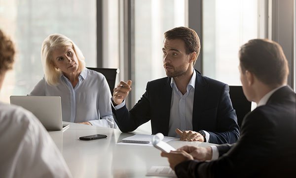 A large group of people sitting in a circle in a conference room, participating in a discussion or presentation. There are banners with DAB logos and a projector screen at the front of the room.