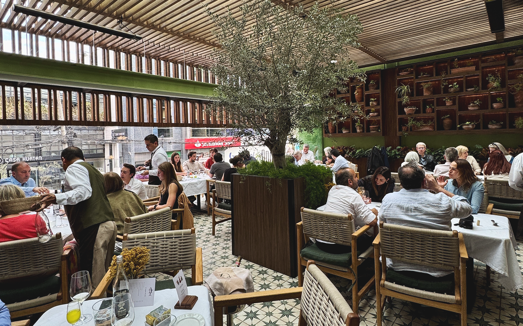 Restaurante con muchas mesas y comensales, grande planta en el centro y paredes decoradas con jarrones de plantas