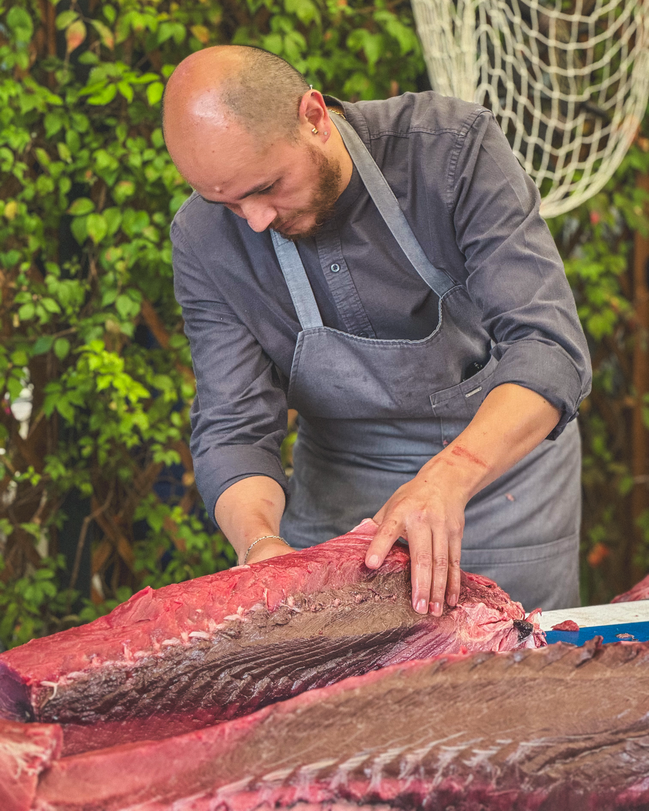 Hombre con delantal negro preparando un pescado en un entorno natural con plantas verdes de fondo.