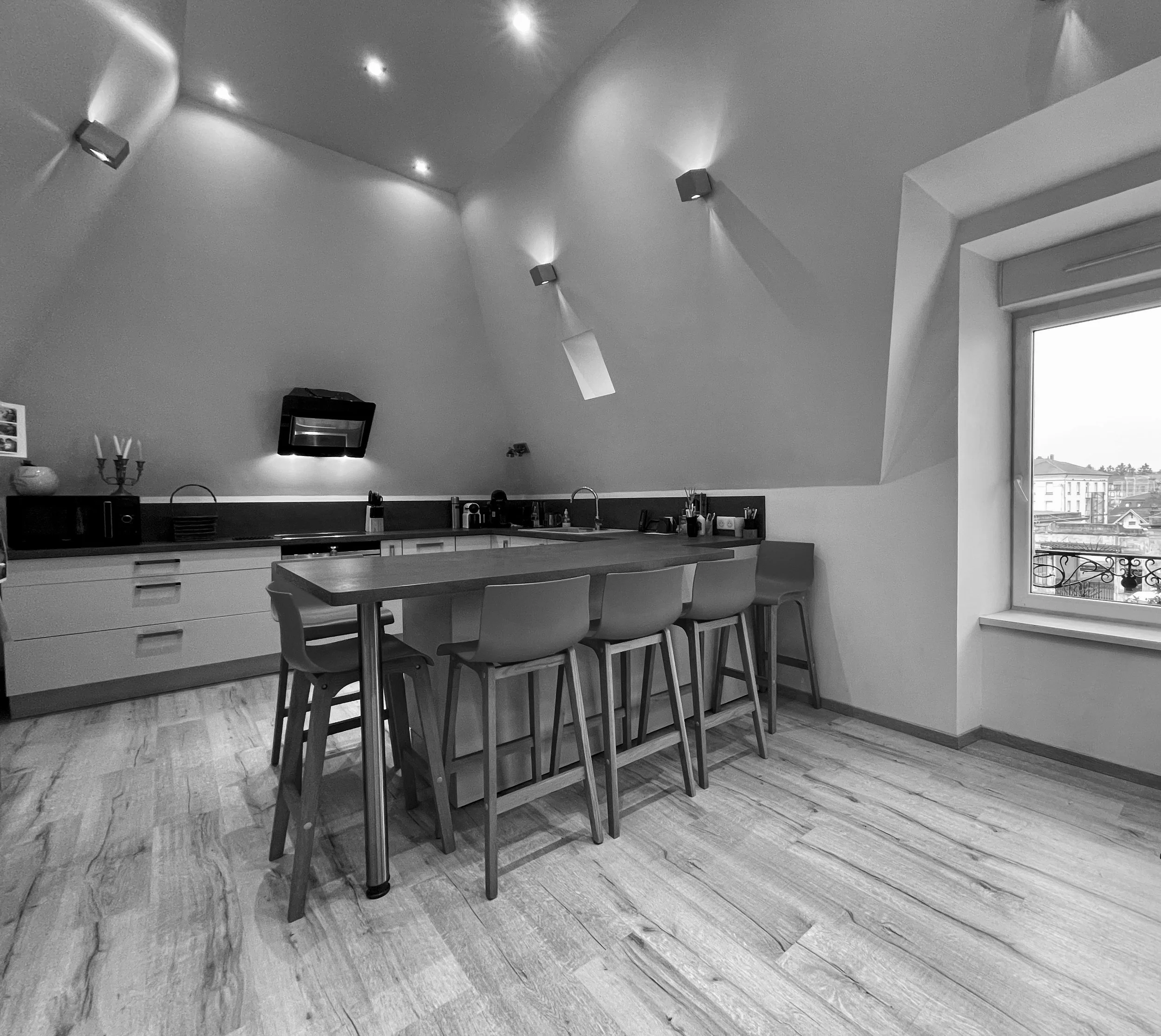 Cuisine moderne avec fenêtre, table de bar avec 5 chaises, armoires blanches, murs et plafond blancs, parquet en bois clair, éclairage encastré au plafond.