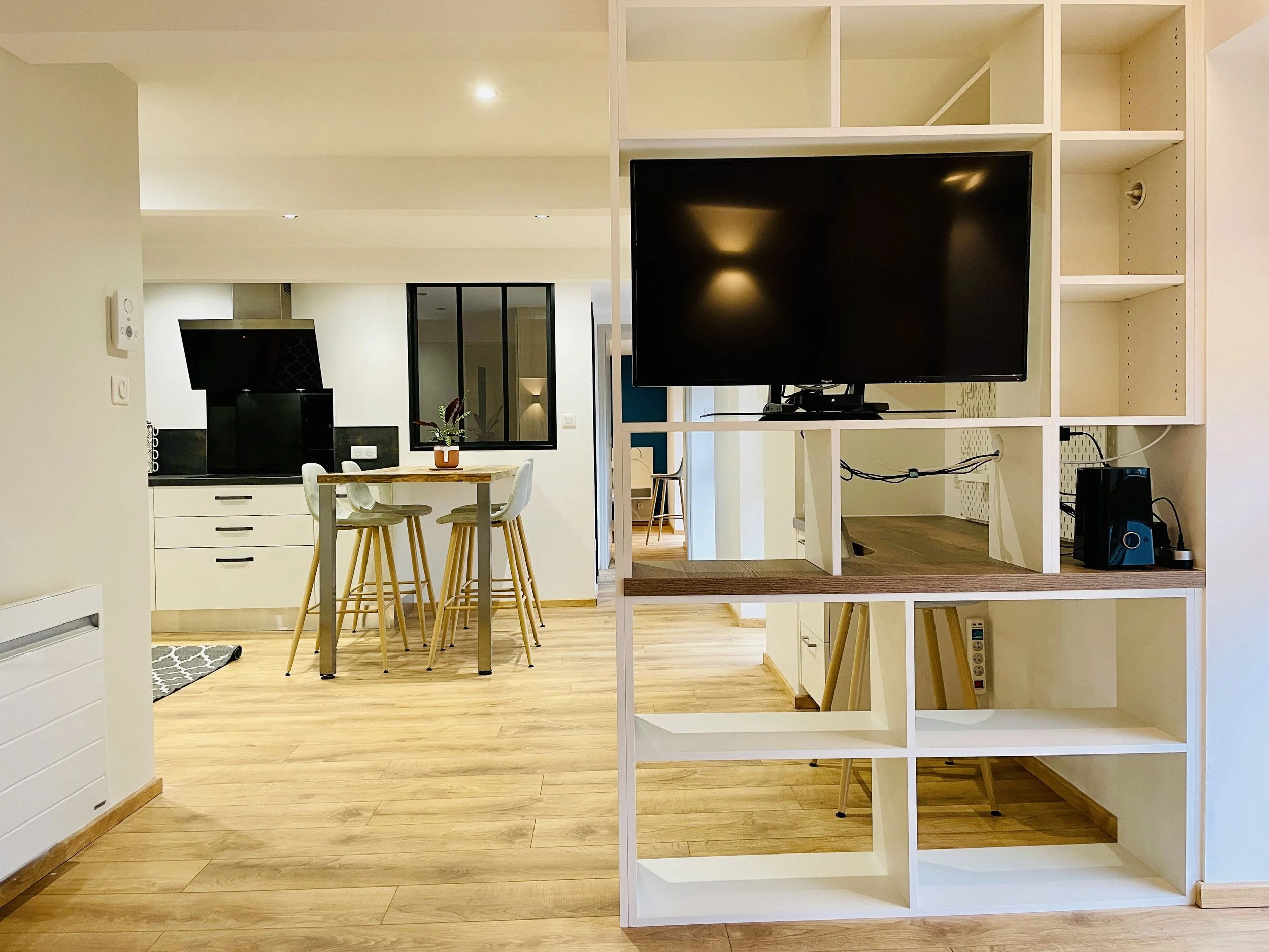 Salon avec meuble de séparation, télévision, cuisine ouverte avec table haute et chaises, sol en bois clair, murs blancs.