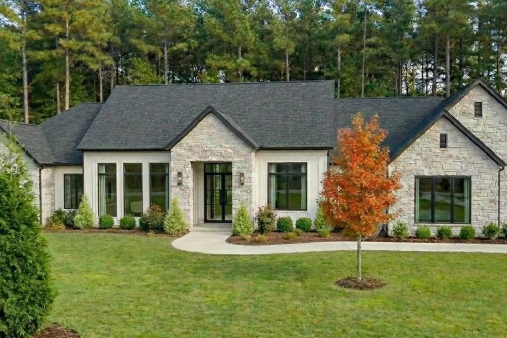 A modern house with stone exterior walls, black window frames, surrounded by a well-maintained lawn with a young orange-leaved tree, and a background of tall trees.