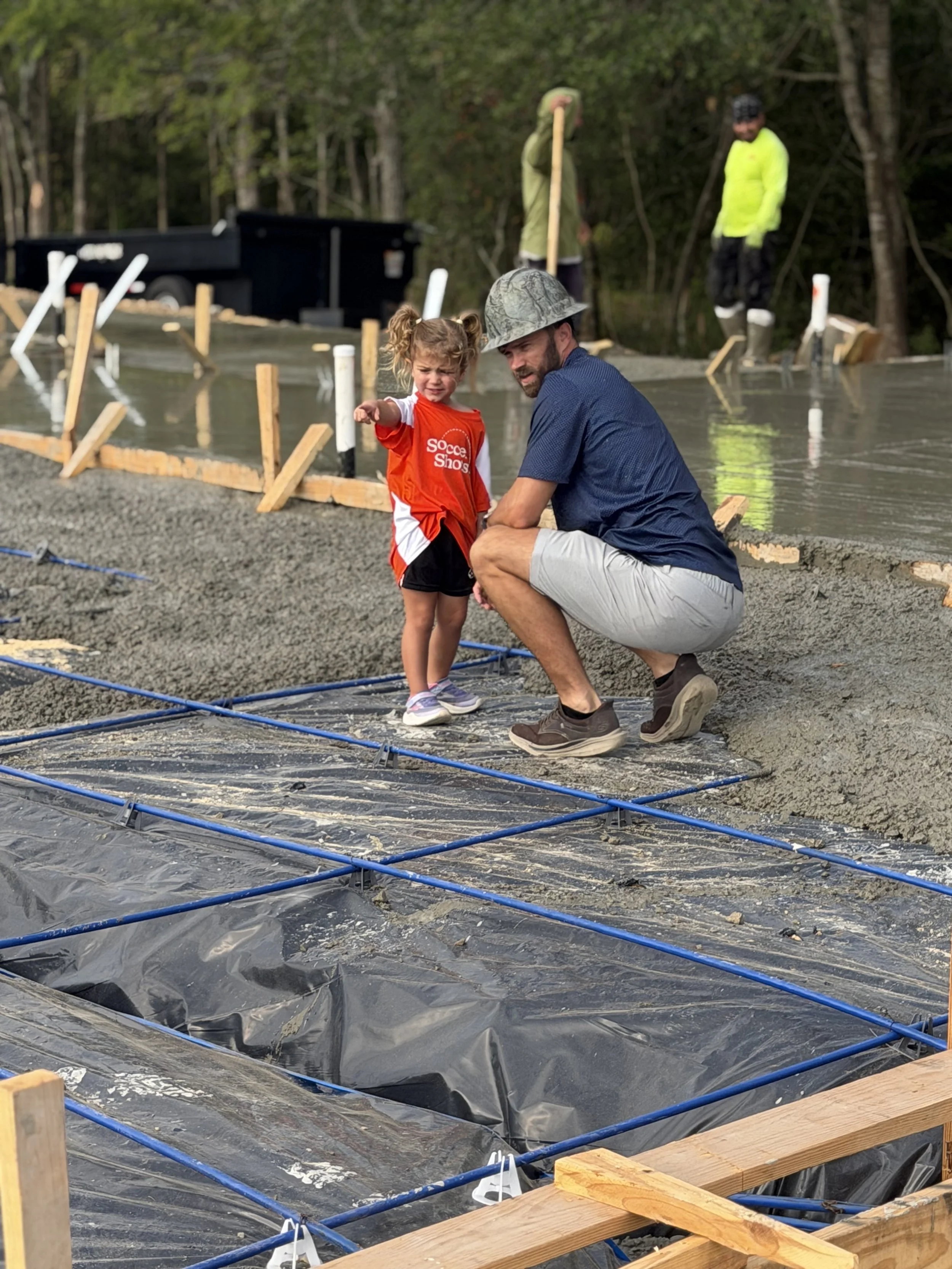 A man and a young girl at a construction site during daytime, with water and black plastic sheeting on the ground, wooden framing, and workers in the background near a tree-lined area.