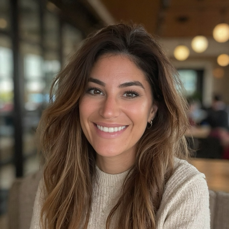 A smiling woman with wavy brown hair, wearing a beige sweater, sitting indoors at a cafe or restaurant.