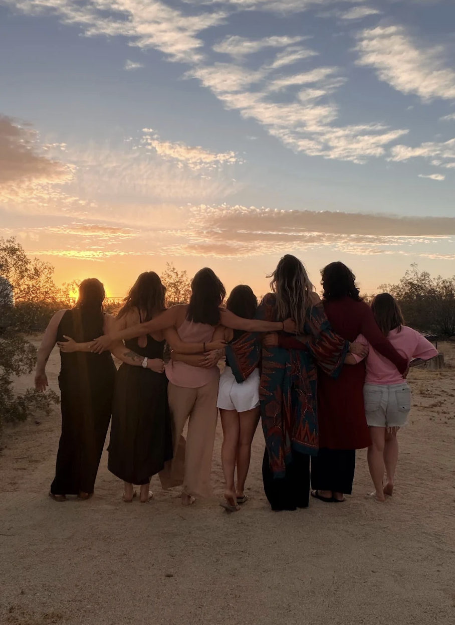 Group of women standing arm-in-arm, watching a sunset after a women's circle while on a retreat.