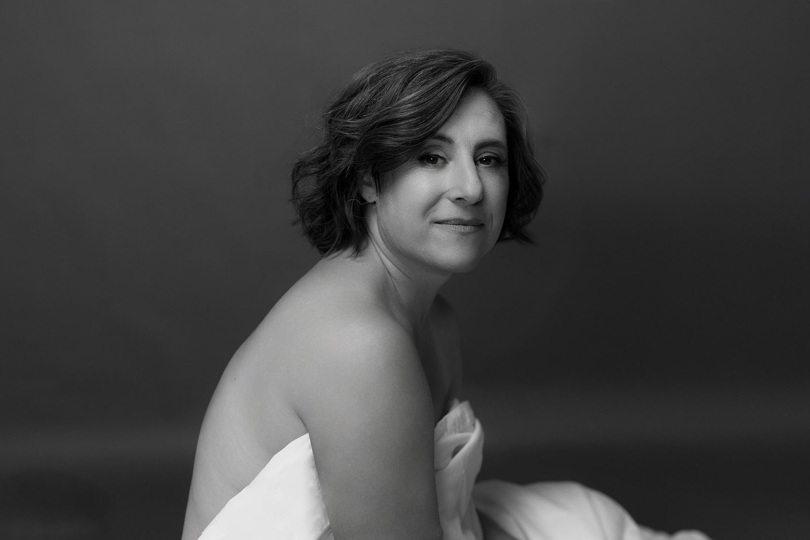 Black and white portrait of a woman with short wavy hair, looking at the camera with a slight smile, draped in a white cloth, against a dark, plain background. Bay Area boudoir and editorial portrait by La Femme Shershow Photography Studio.