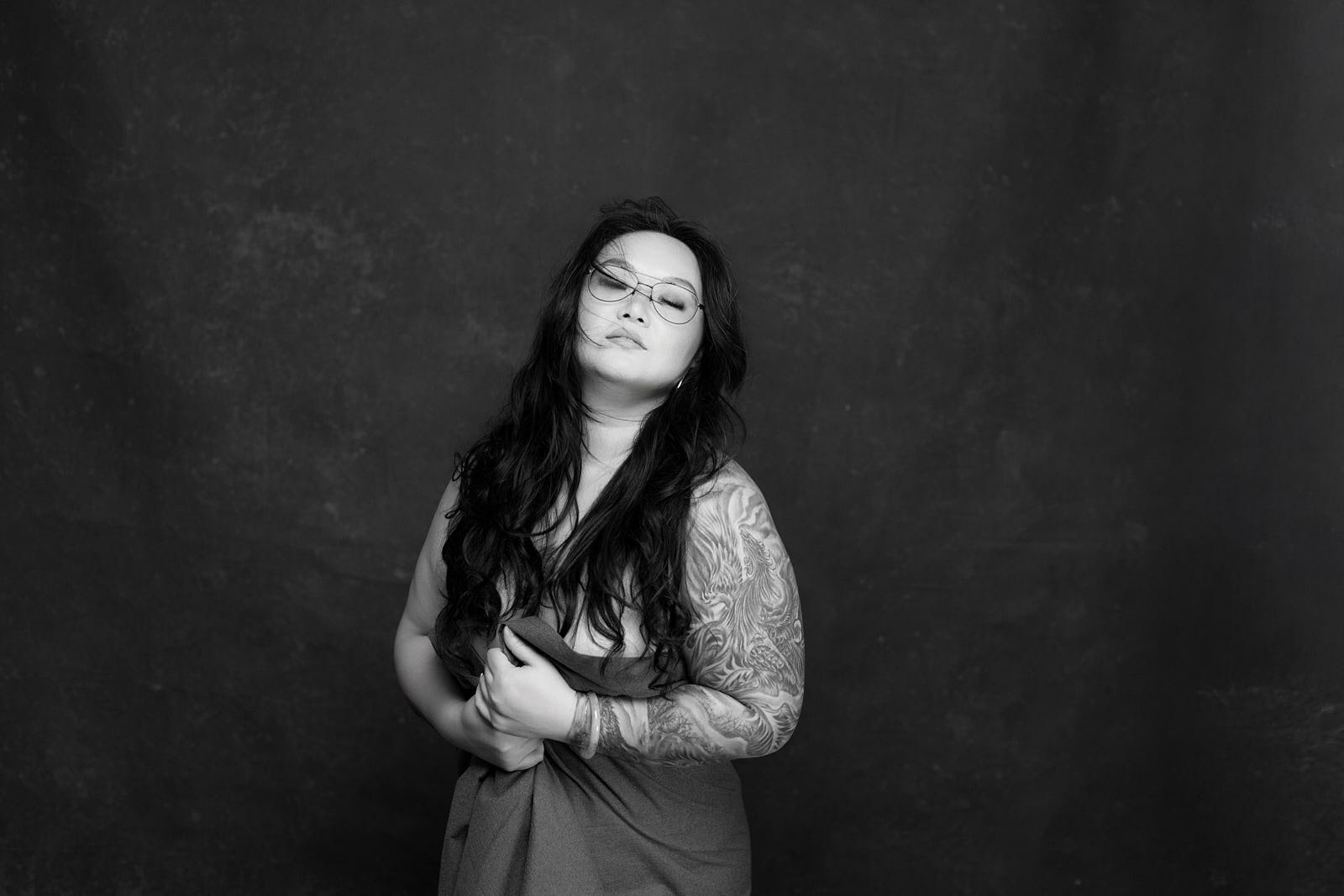 Black and white portrait of a woman with long dark hair wearing glasses, holding her dress with one hand, standing against a dark background. Bay Area boudoir and editorial portrait by La Femme Shershow Photography Studio.