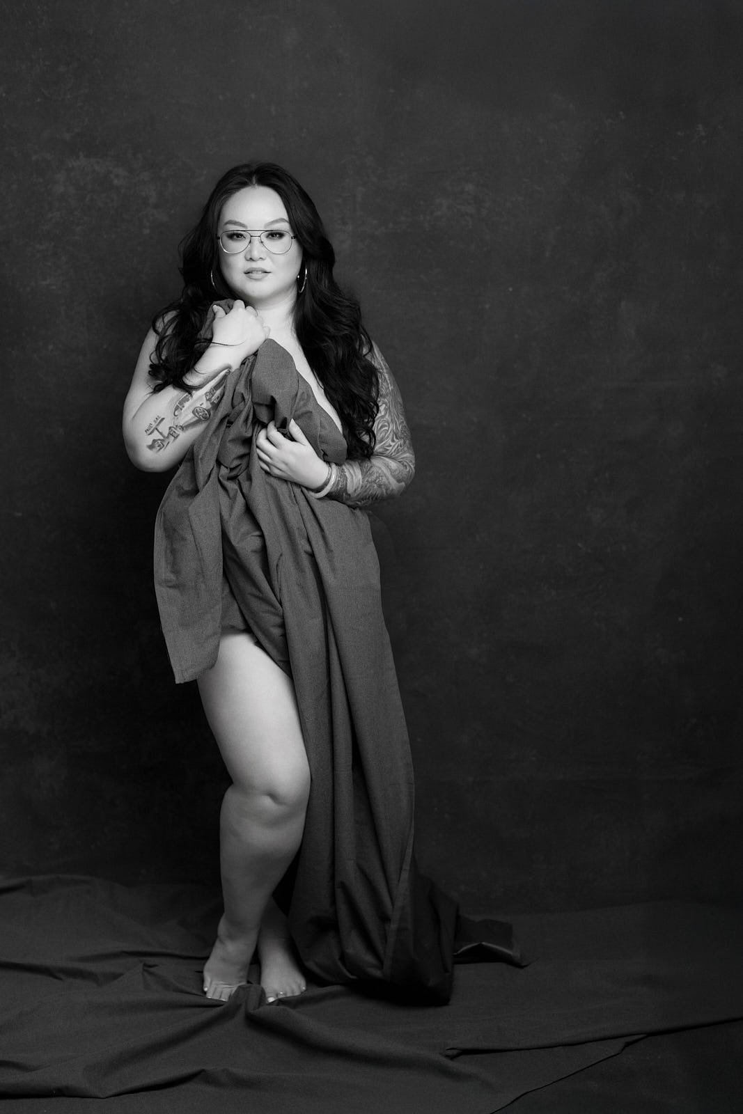 Black and white portrait of a woman with long dark hair, glasses, and tattoos, holding fabric, standing barefoot against a dark textured background. Bay Area boudoir and editorial portrait by La Femme Shershow Photography Studio.