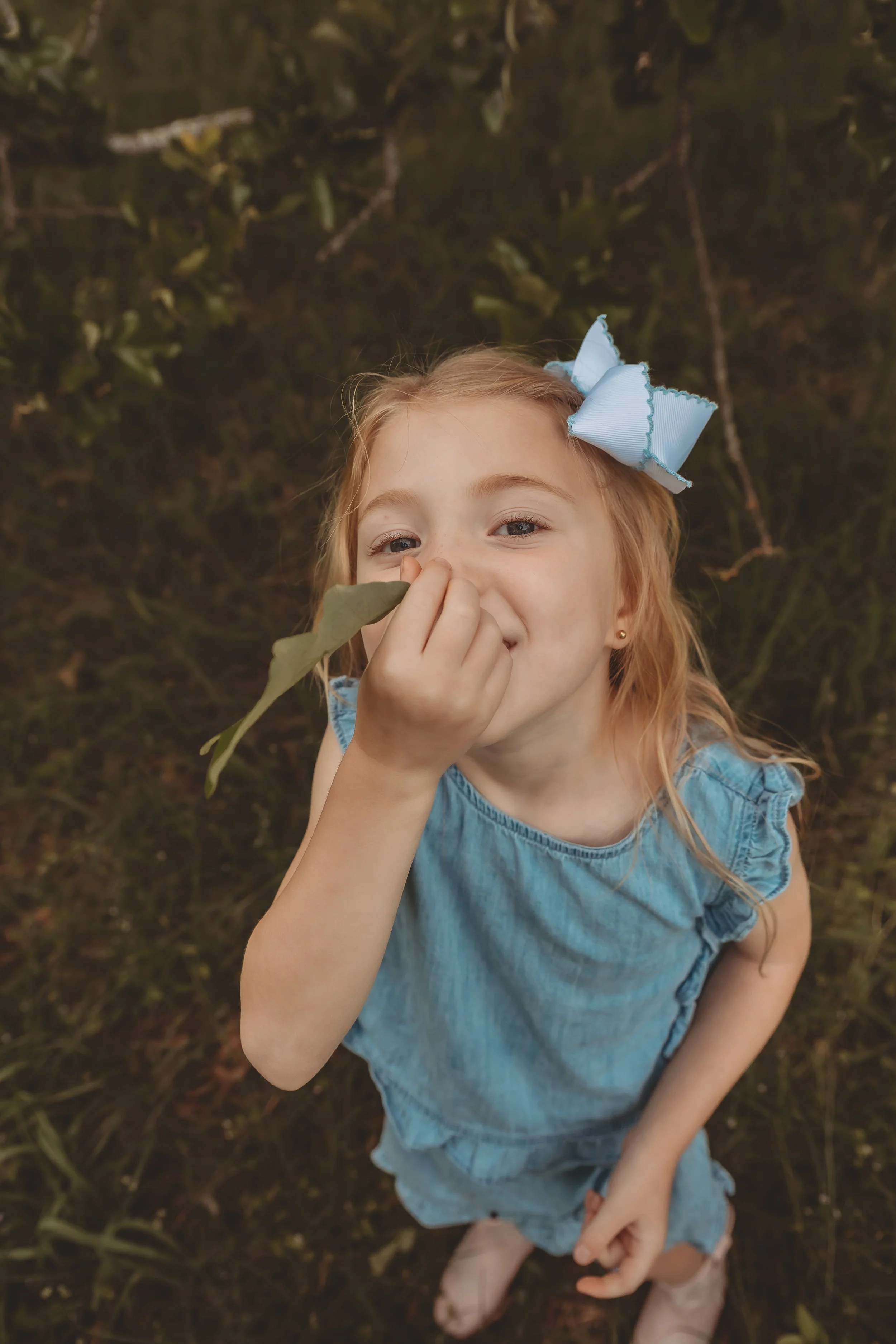 A young girl with red hair and a blue dress standing outdoors on grass, holding a leaf close to her nose, and wearing a large blue bow in her hair.