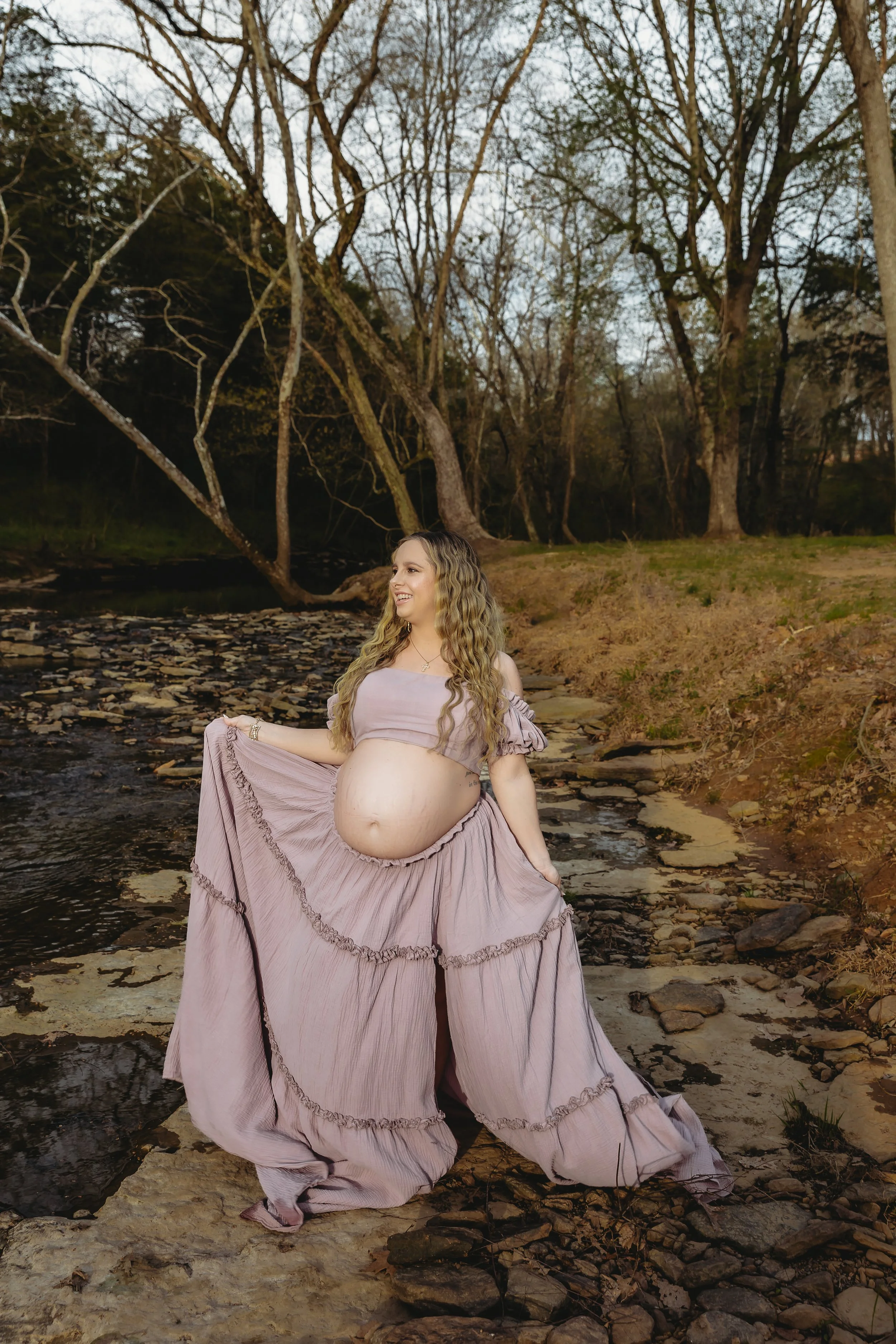 A pregnant woman with long curly hair stands outdoors on a rocky riverbank during dusk, wearing a light purple flowing dress, smiling and looking to the side with trees in the background. Maternity Session. Client Closet.