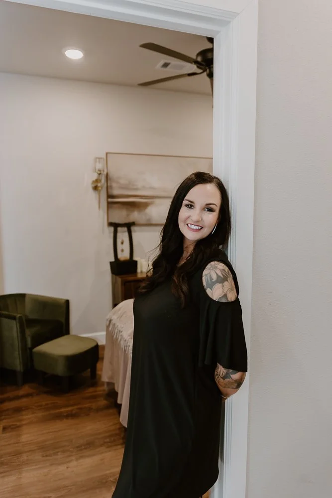 A woman with dark hair and tattoos, dressed in black, standing in a doorway and smiling.