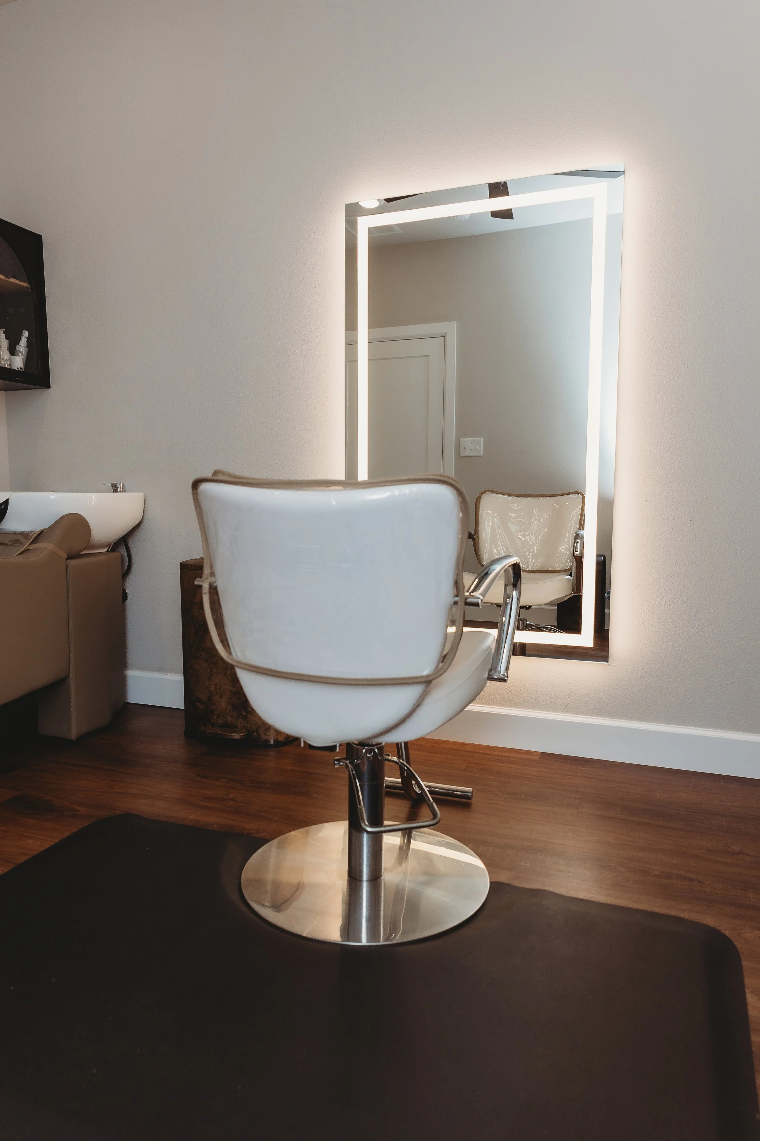 A styling station in a salon with a white salon chair, a full-length mirror with LED lighting, and a brown wooden floor. There is a white wall in the background and some salon equipment visible.