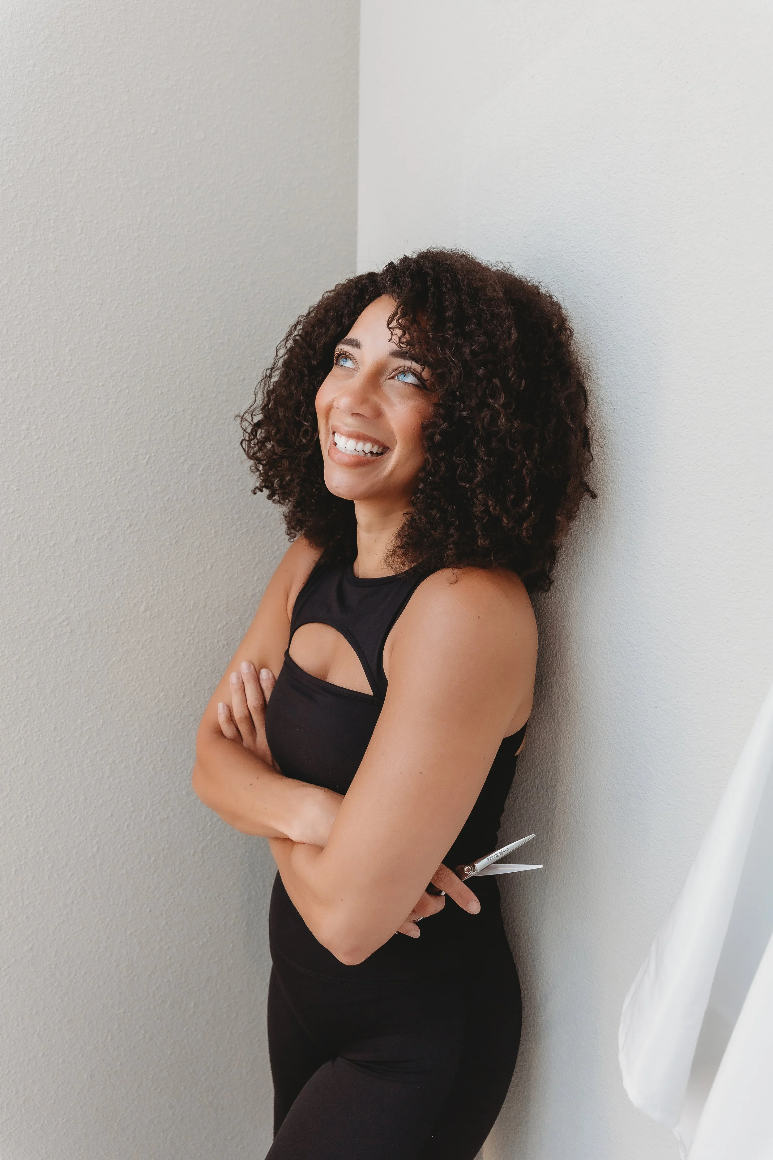 A woman with curly hair leaning against a wall, smiling and looking up. She is dressed in a black sleeveless top and black pants, standing with her arms crossed. A white curtain is visible on the right side of the image.