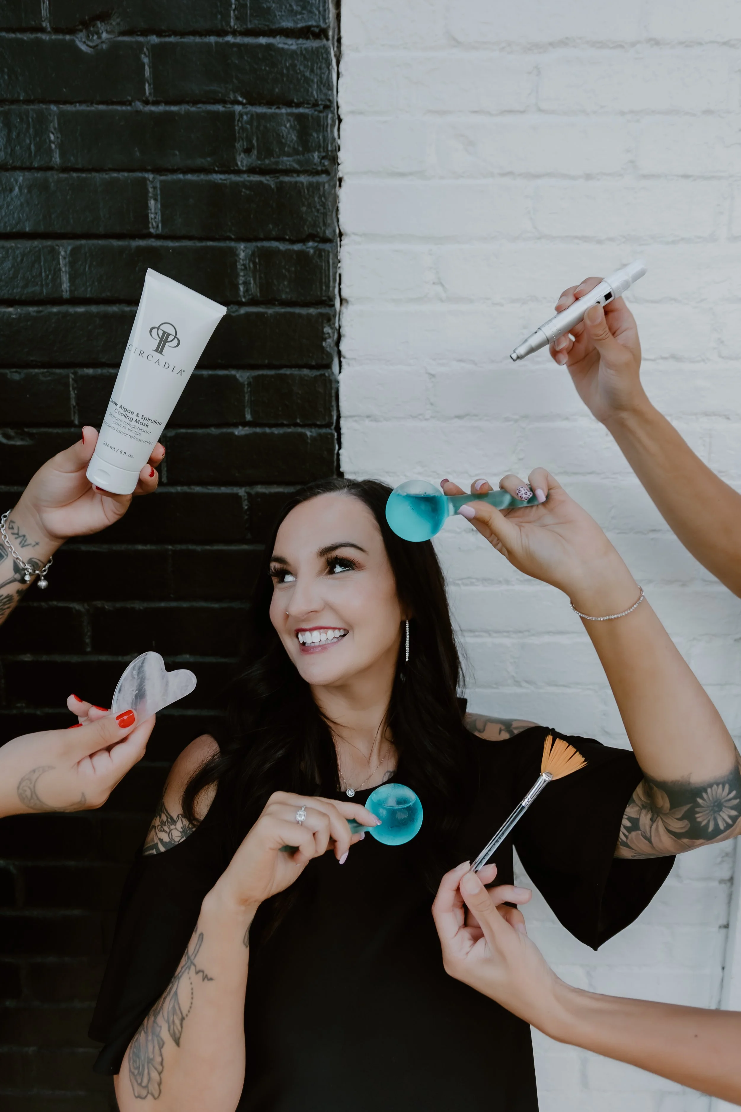 A smiling woman with dark hair and tattoos getting a beauty treatment with facial tools and skincare products against a black and white brick wall.