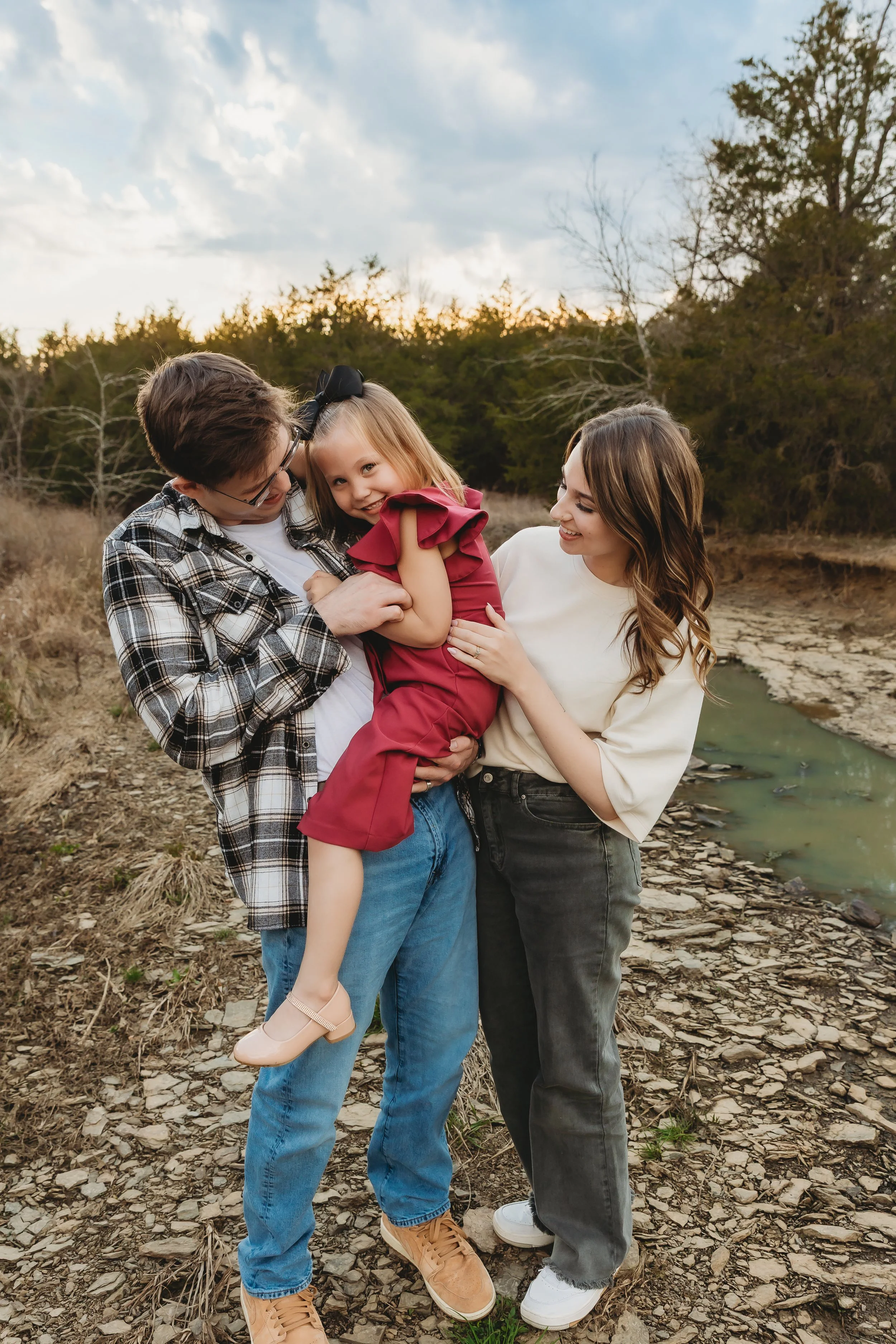 A family of three standing outdoors near a small stream with trees and a cloudy sky in the background. The father is holding a young girl, and the mother is standing next to them, all smiling and looking at each other.
