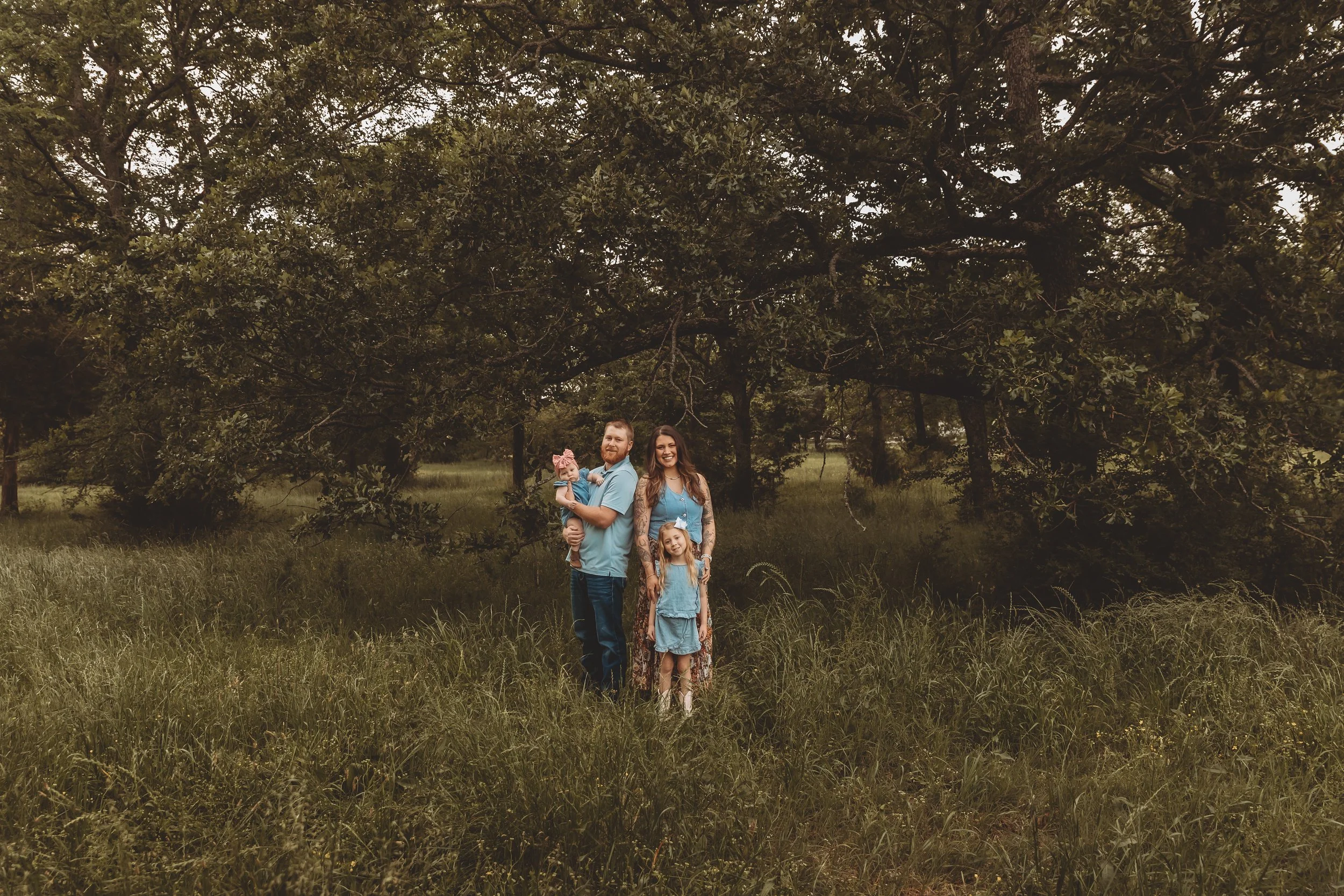 A family of four standing in a grassy clearing with large trees overhead. The man is holding a baby girl, and a young girl is standing in front of the woman, who has tattoos on her arms. All are smiling and wearing casual clothing.