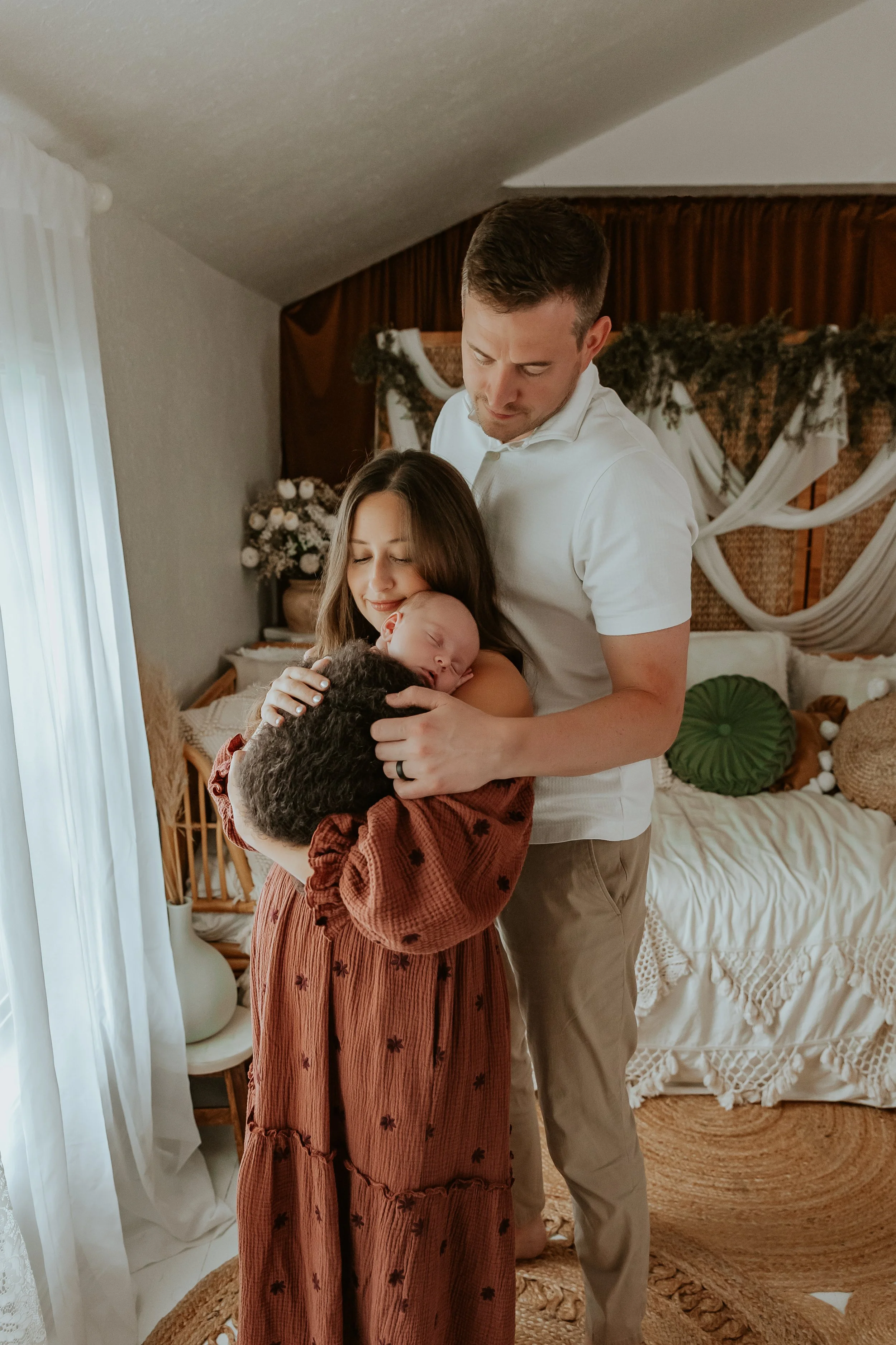 A man and a woman with a newborn baby in a cozy, decorated bedroom, with the woman holding the baby and the man leaning in close, all smiling softly. Newborn Studio Session