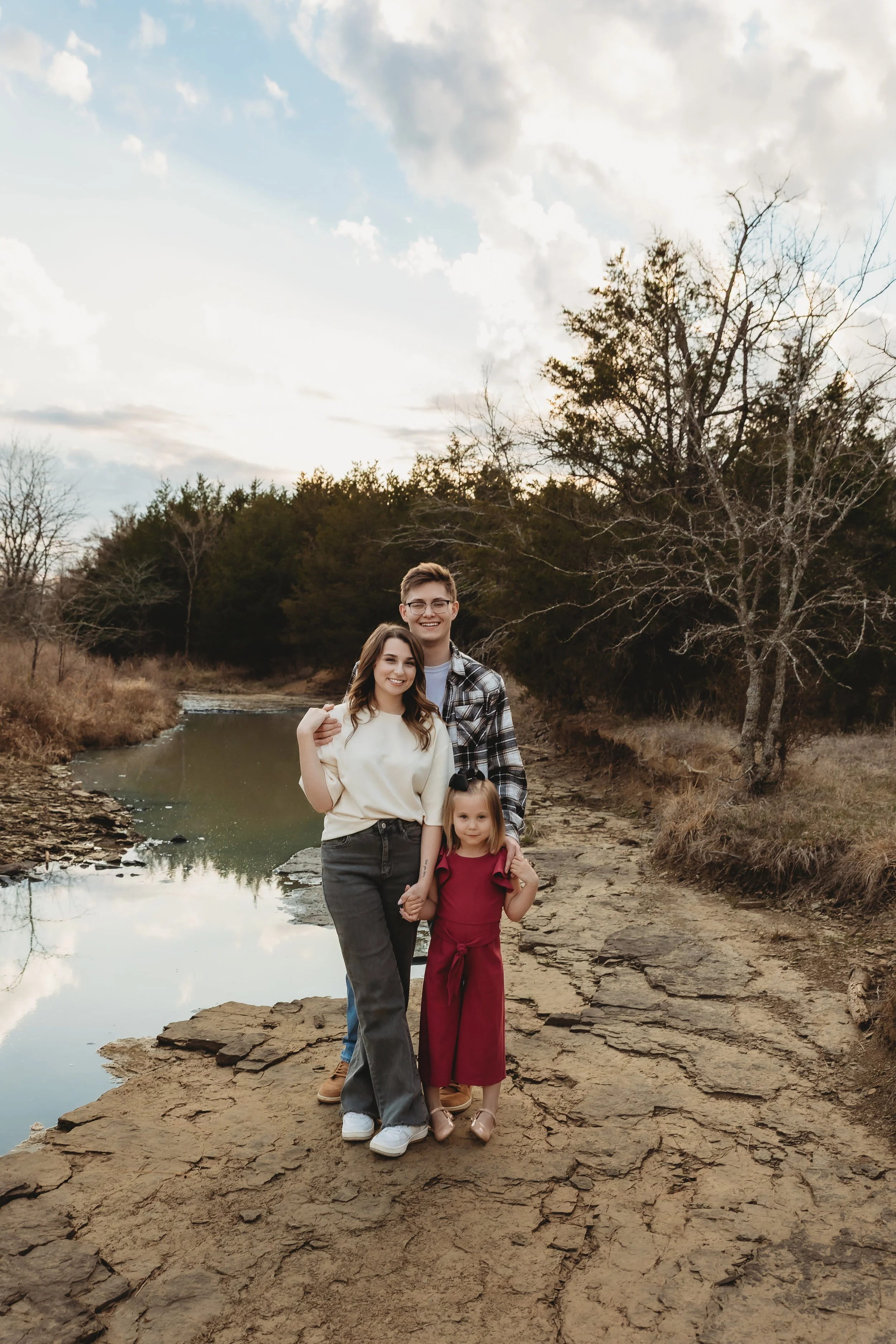 A family of four stands on a rocky riverbank amid leafless trees, with a cloudy sky overhead, posing for a photo during a hike or outdoor trip.