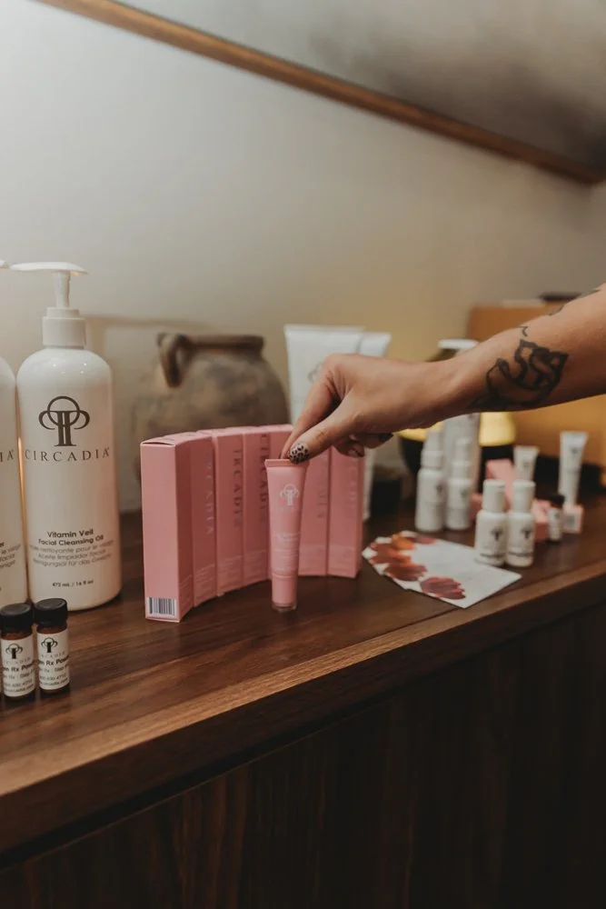 A person with a tattoo on their arm is holding a pink skincare product tube on a wooden surface, surrounded by skincare bottles, boxes, and sample vials.
