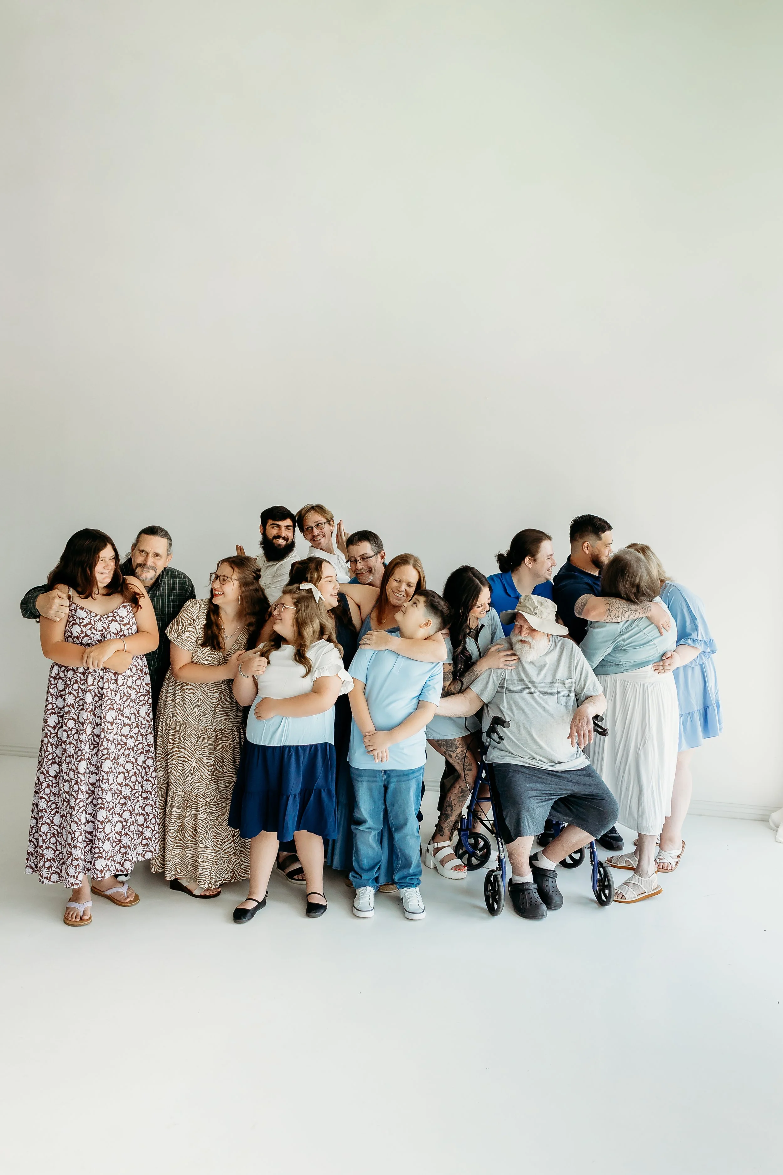 A large group of diverse people, including children, adults, and elderly individuals, gathered closely together against a plain white background. Some are smiling and embracing, creating a joyful and inclusive atmosphere.