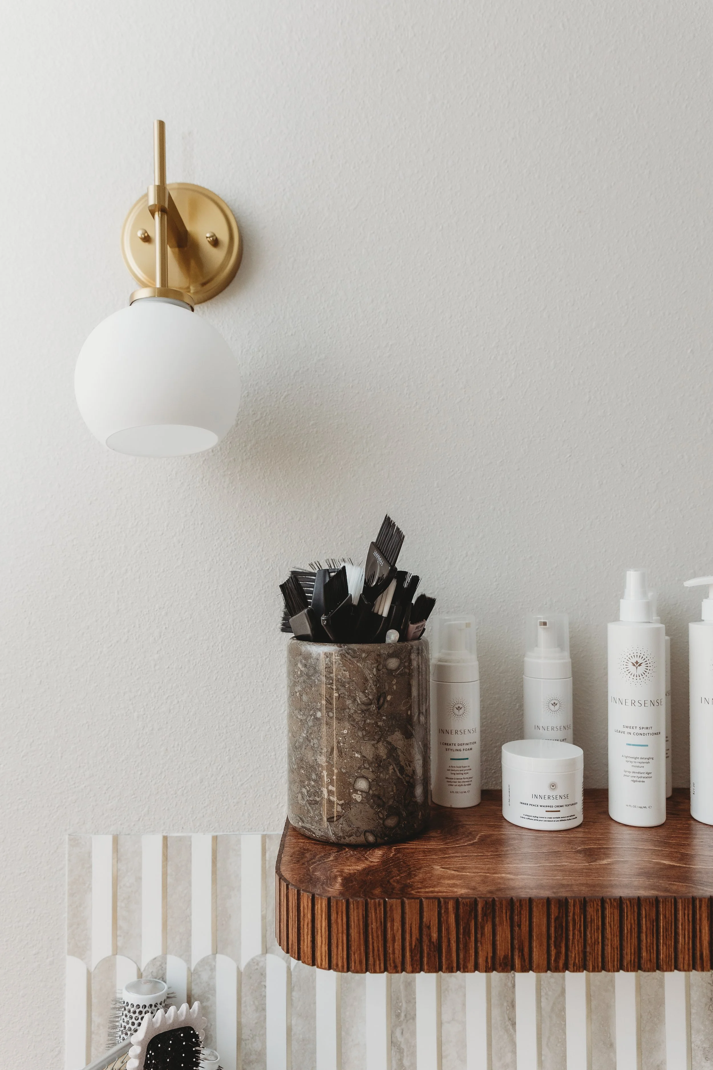 Bathroom or vanity area with a brass wall-mounted light fixture, a marble container holding hairbrushes or styling tools, and several bottles of InnerSense hair products on a wooden surface.