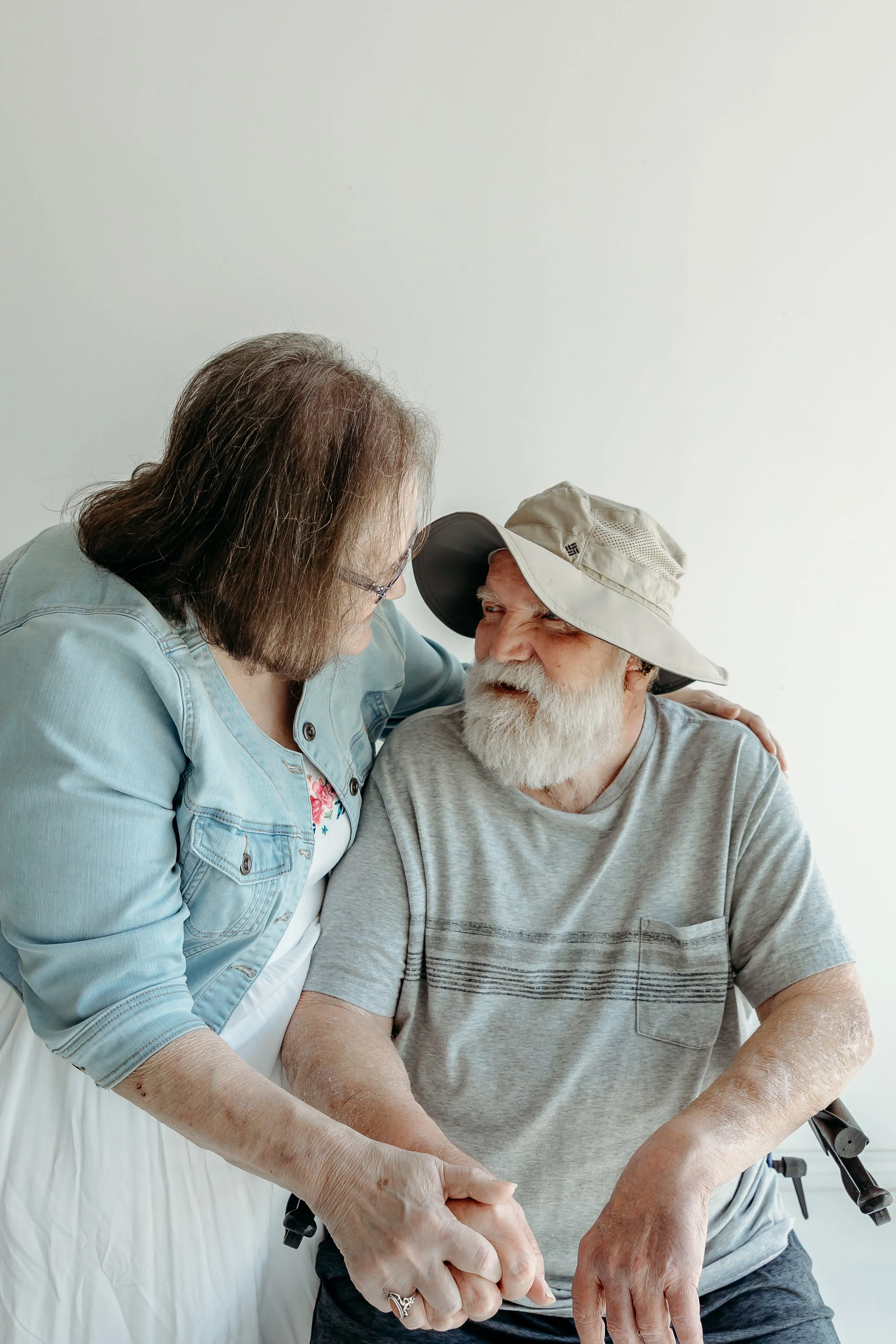 A woman and an elderly man are holding hands, smiling and looking at each other in a room with a plain white wall.