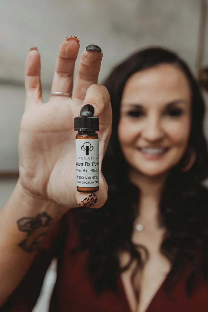 Woman with dark hair smiling, holding a small amber bottle labeled 'Oxygen Rx Powder' close to the camera.