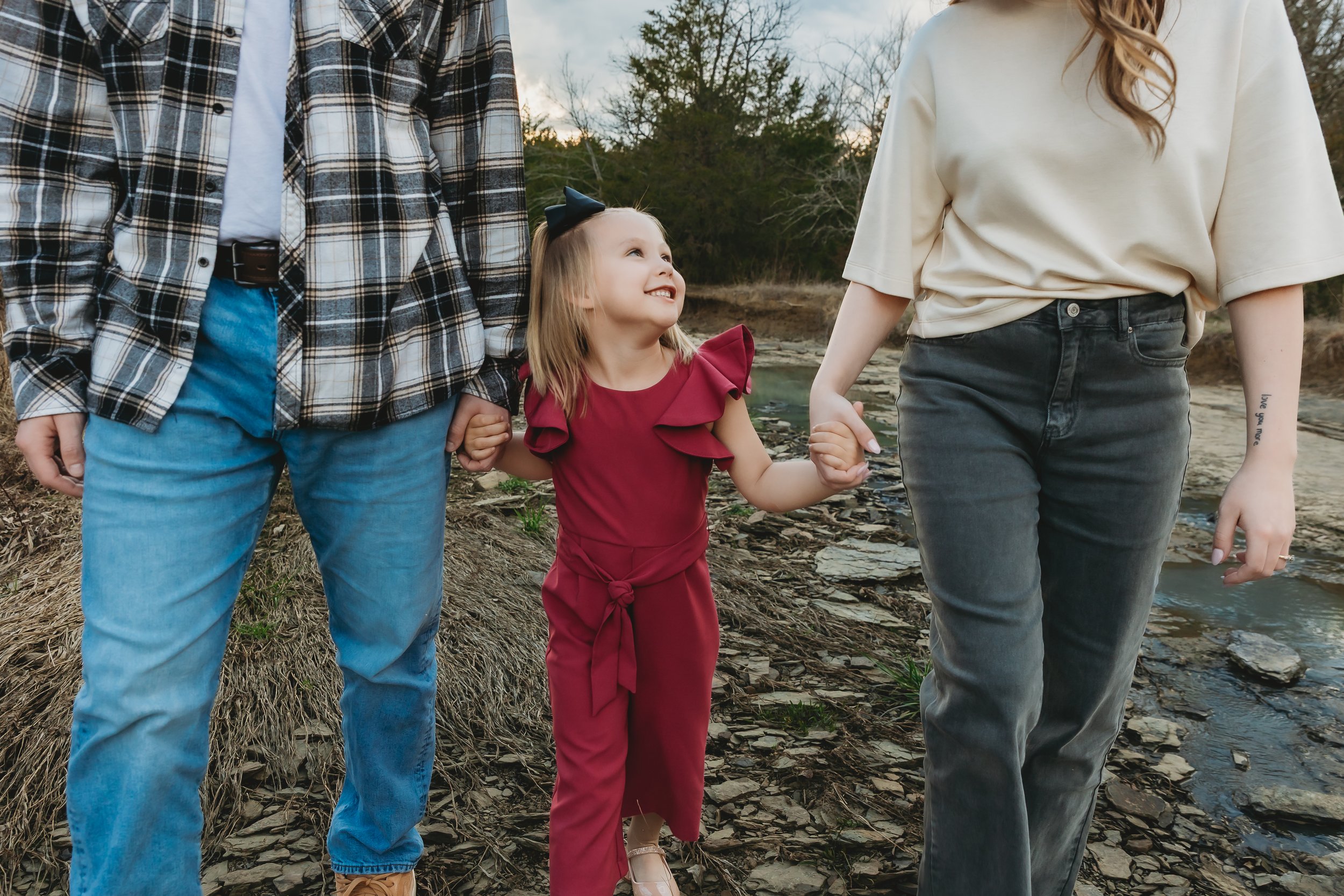 A family walking outdoors near a body of water during daytime. The image shows a child holding hands with two adults, likely her parents, on a natural, rocky shoreline with trees in the background.