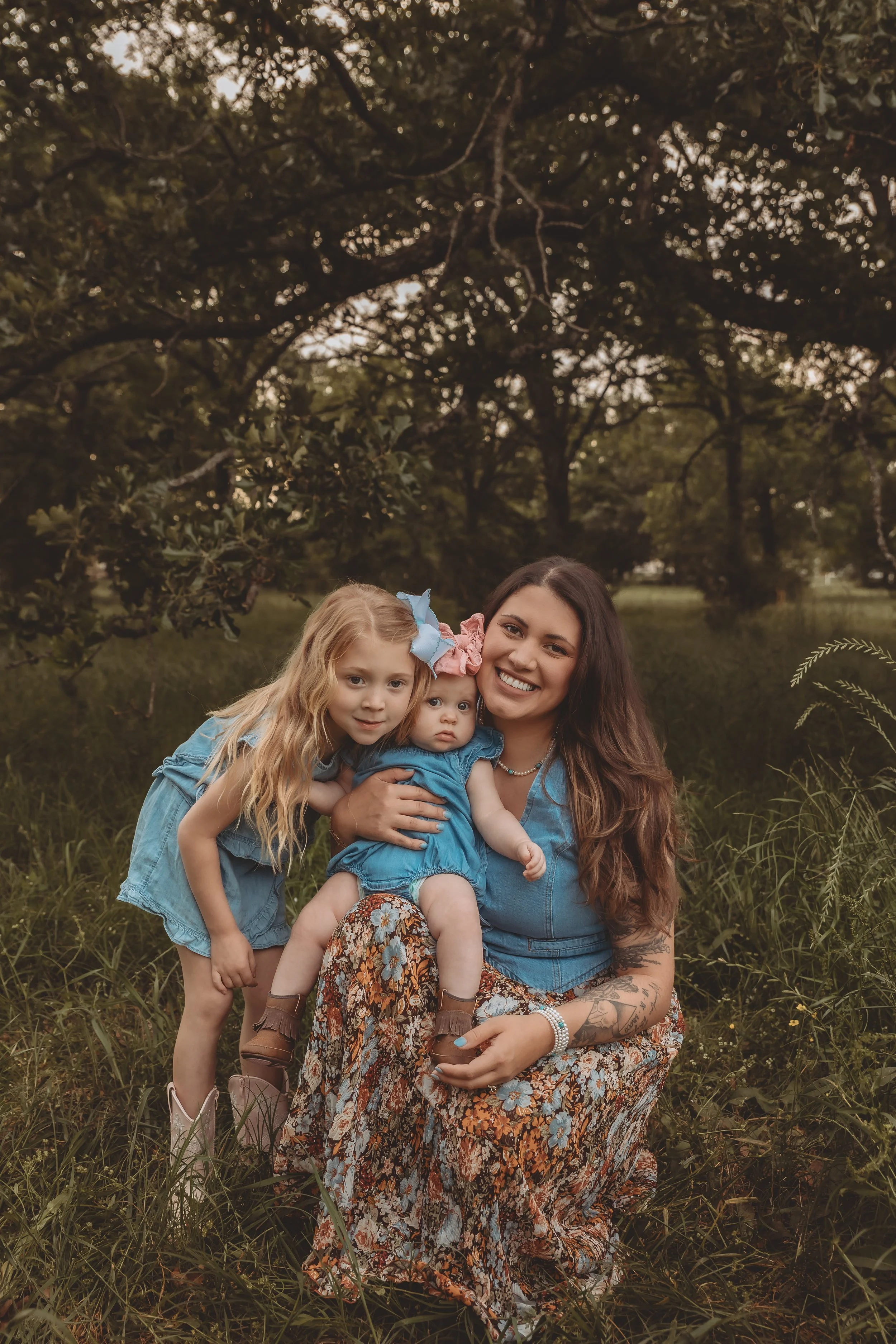 A woman with long brown hair and tattoos, wearing a floral skirt and denim top, is sitting outdoors in grass with two young girls, one with long blonde hair and a blue bow, the other with short brown hair and a pink bow, all smiling and dressed in bl
