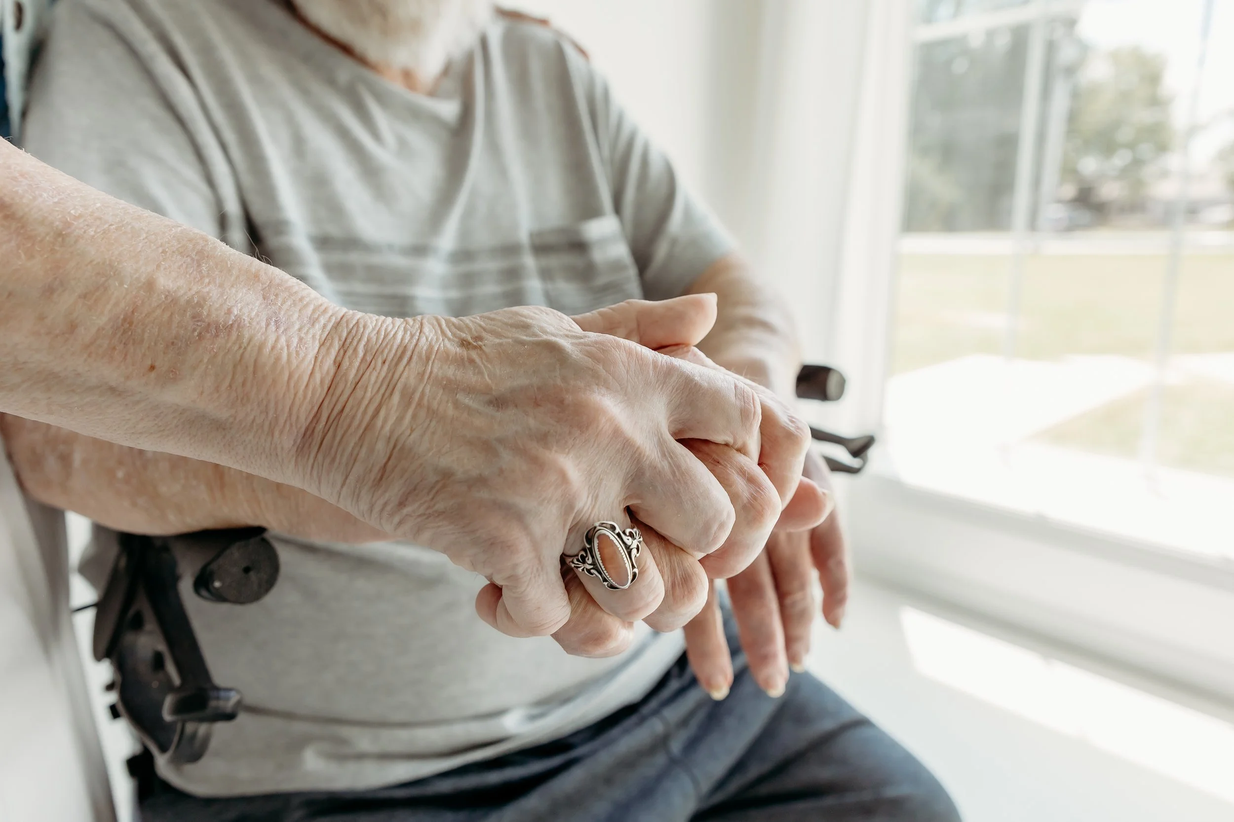 Close-up of elderly person's hands resting on their lap, with visible wrinkles and a silver ring, in front of a window.